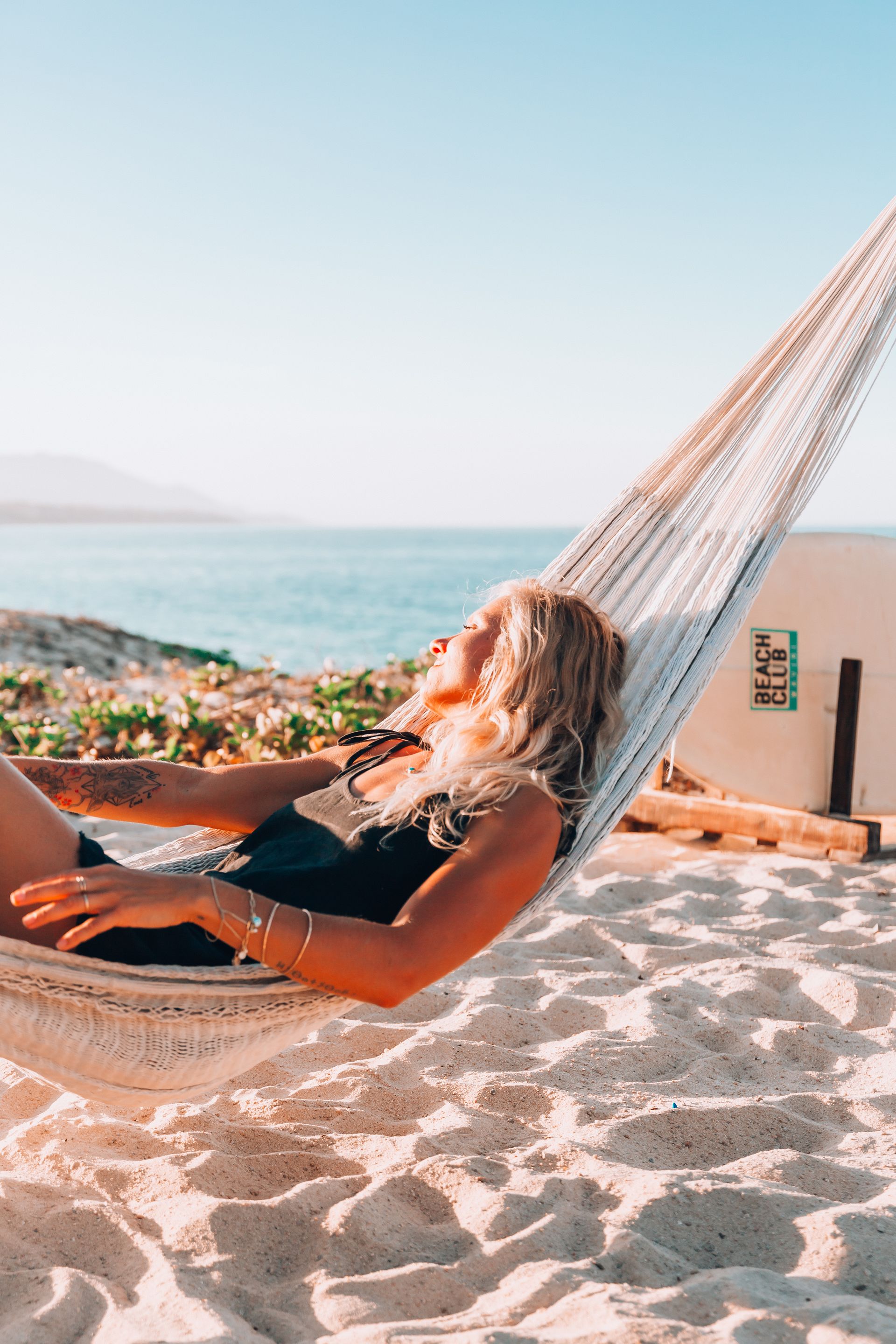 Woman relaxing in a hammock on a sandy beach, sunny day, ocean in the background.