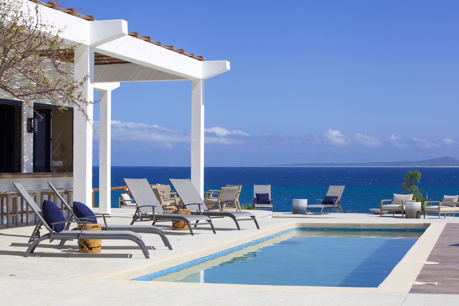 Poolside lounge chairs overlooking the ocean, next to a white villa with a pergola.