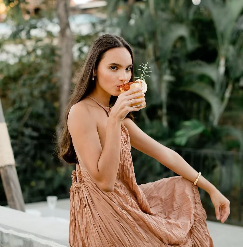 Woman in a peach-colored dress sips a cocktail with a garnish. Outdoors, lush greenery in the background.