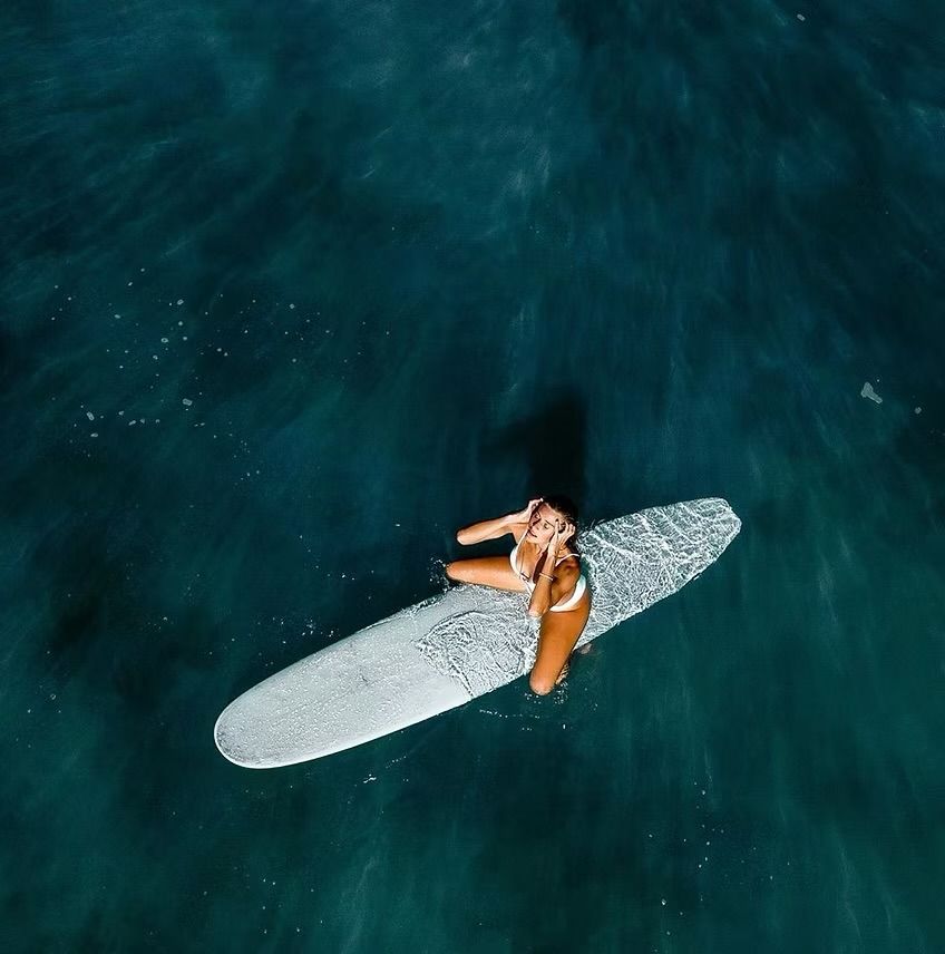 Woman in white bikini sits on a surfboard in blue ocean water, looking up.