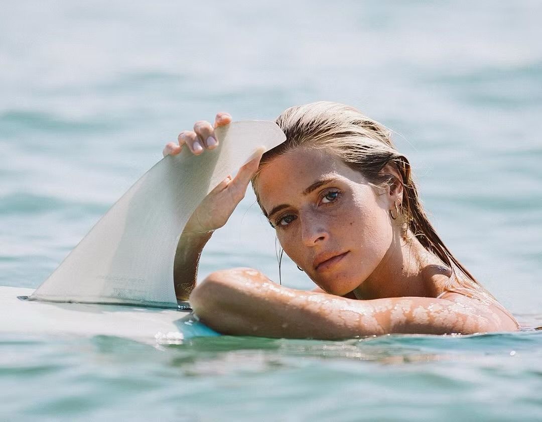 Woman in the ocean, playfully holding a shark fin-shaped board, looking toward the camera. Blue water, sunny.