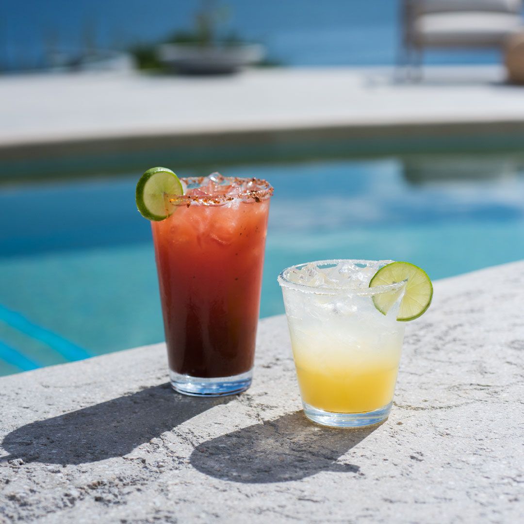 Two cocktails, one red and one yellow, sit by a pool with lime garnishes.