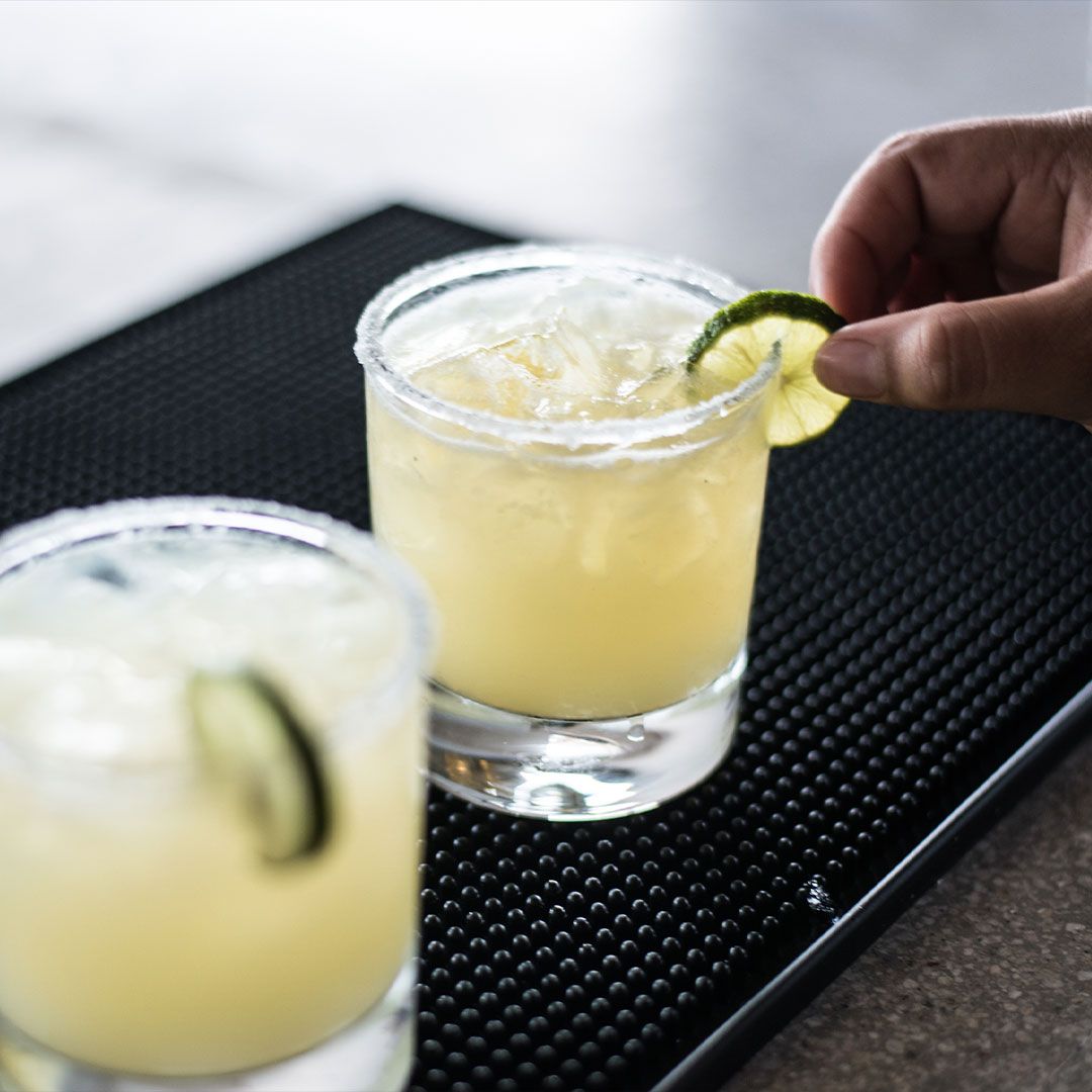 Two margaritas, one with lime garnish being added by hand, on a black bar mat.