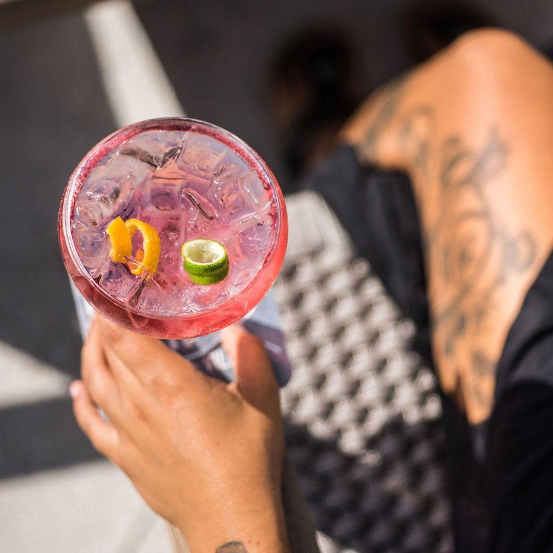 Person holding a pink cocktail with ice, garnished with lime and orange peel.