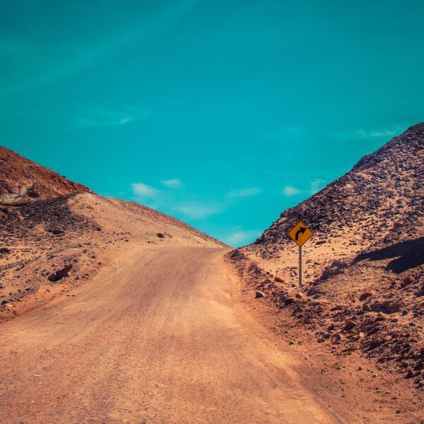 Dirt road winding between brown hills, blue sky, yellow curve sign.