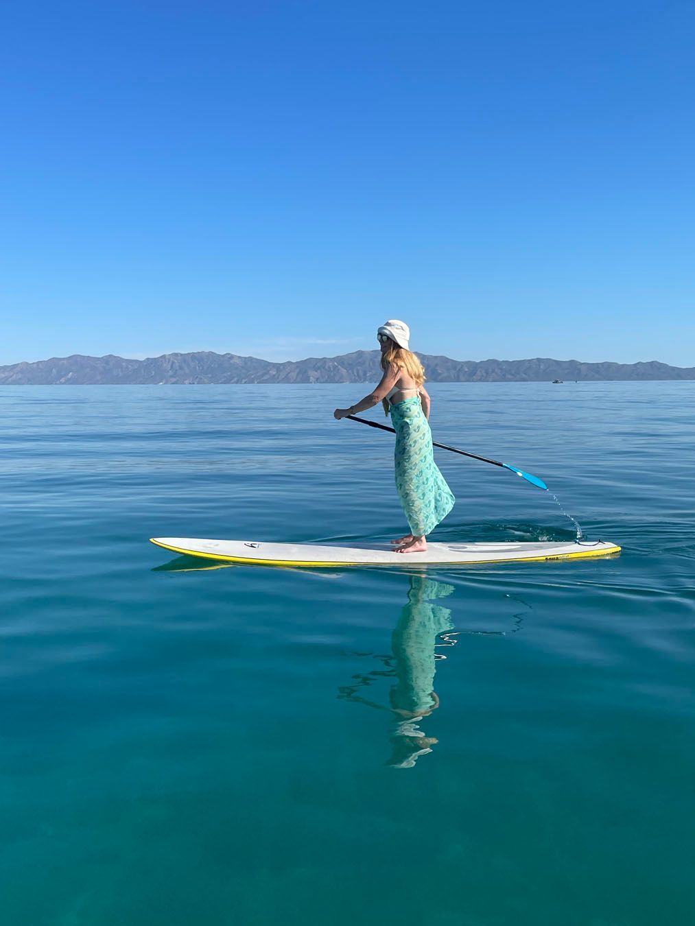 Woman paddleboards on a calm, turquoise sea with mountains in the distance under a clear blue sky.