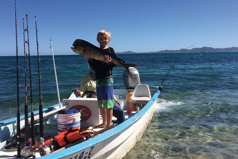 Boy holds a fish on a boat in sunny, blue water near the shore. Fishing rods are visible.