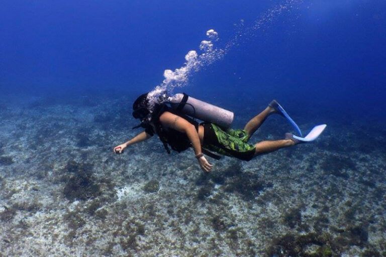 Scuba diver swimming underwater, exhaling bubbles; wearing fins, black vest, and green shorts.