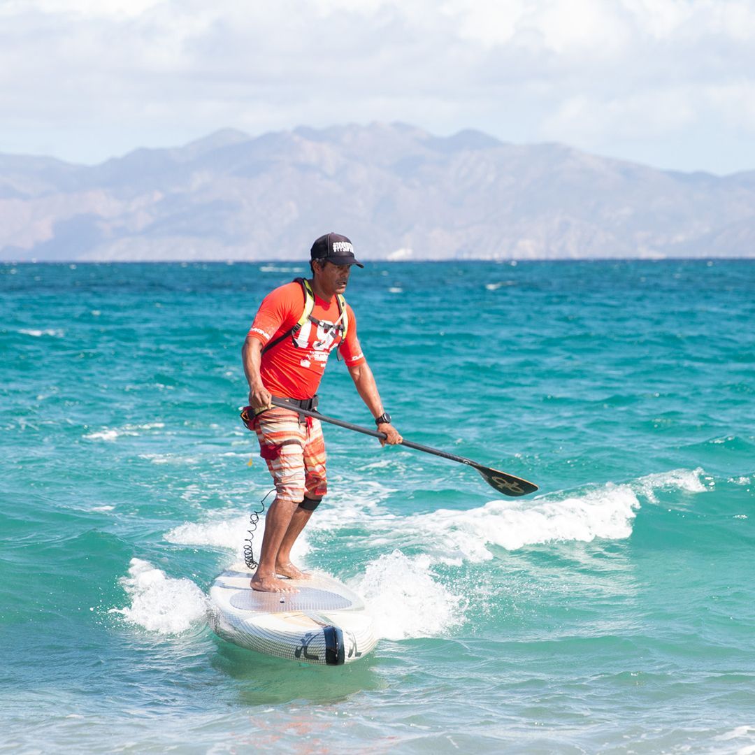 Man paddleboarding on a wave in front of mountains. He wears a red shirt, shorts, and a hat. Turquoise water.