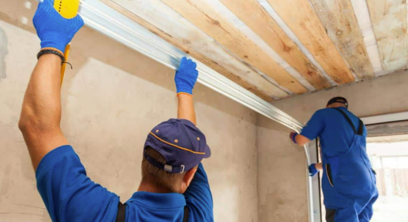 Two workers in blue uniforms install a metal track for a garage door on a bare, unfinished concrete ceiling and wall.