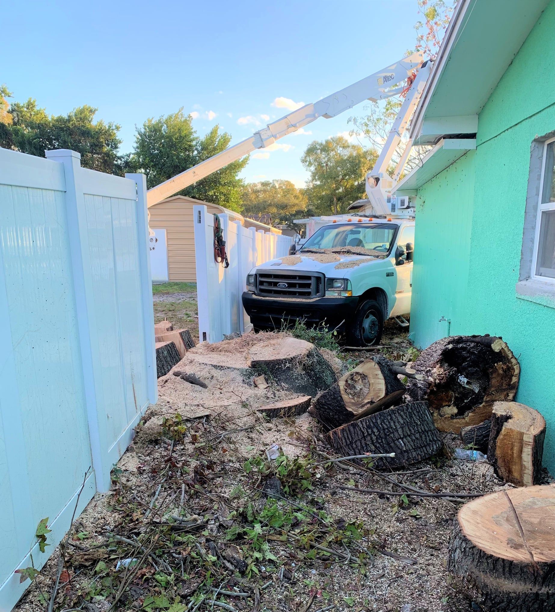 Trucks crushed a fence and trailer beside a turquoise house, with debris scattered in the yard.