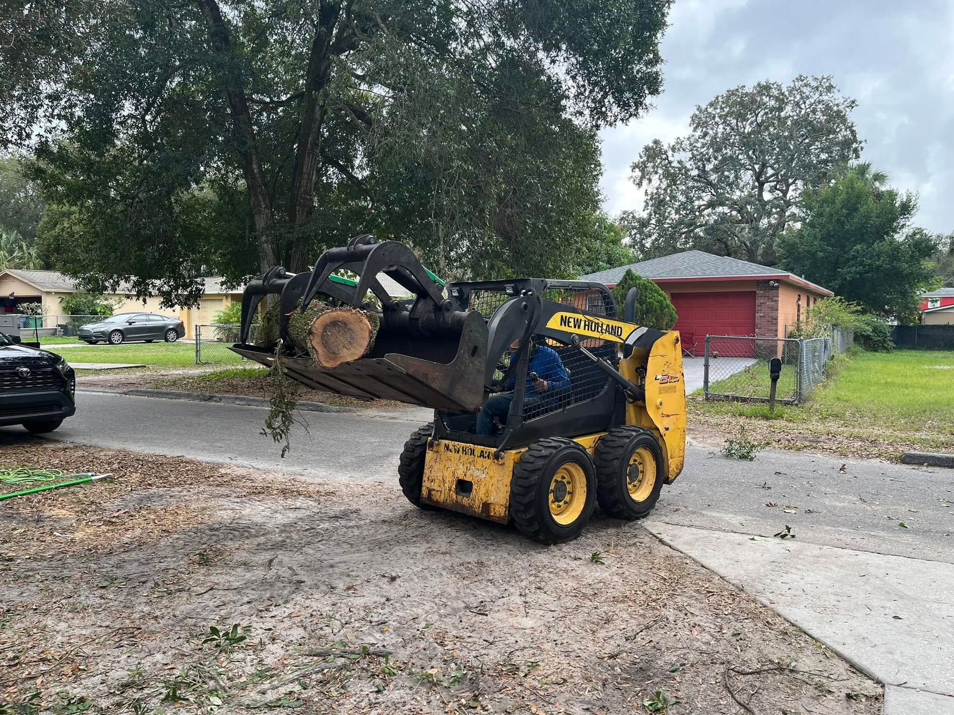 Yellow skid-steer loader lifting a large dirt-filled root ball beside a street and driveway in a residential yard