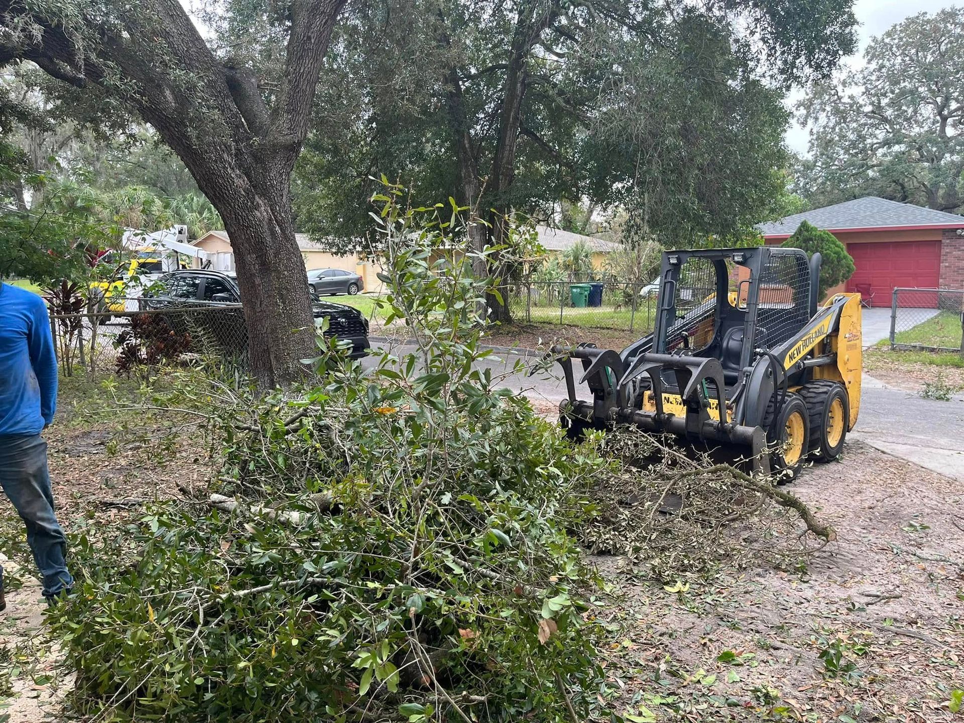 Backhoe loader clearing brush beside a large tree and gravel driveway near a red building