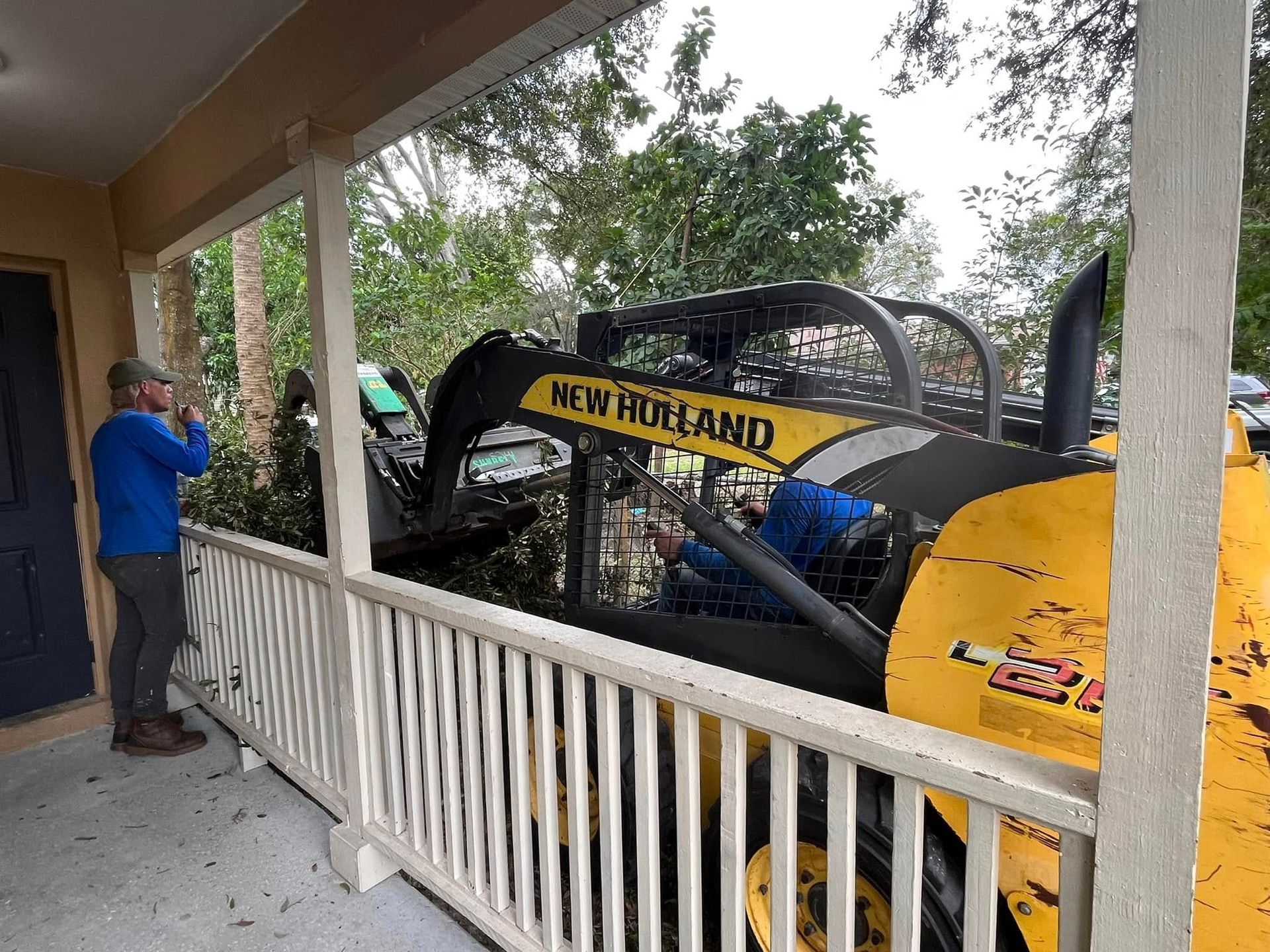 Man in blue shirt beside New Holland construction vehicle on a porch in a wooded area
