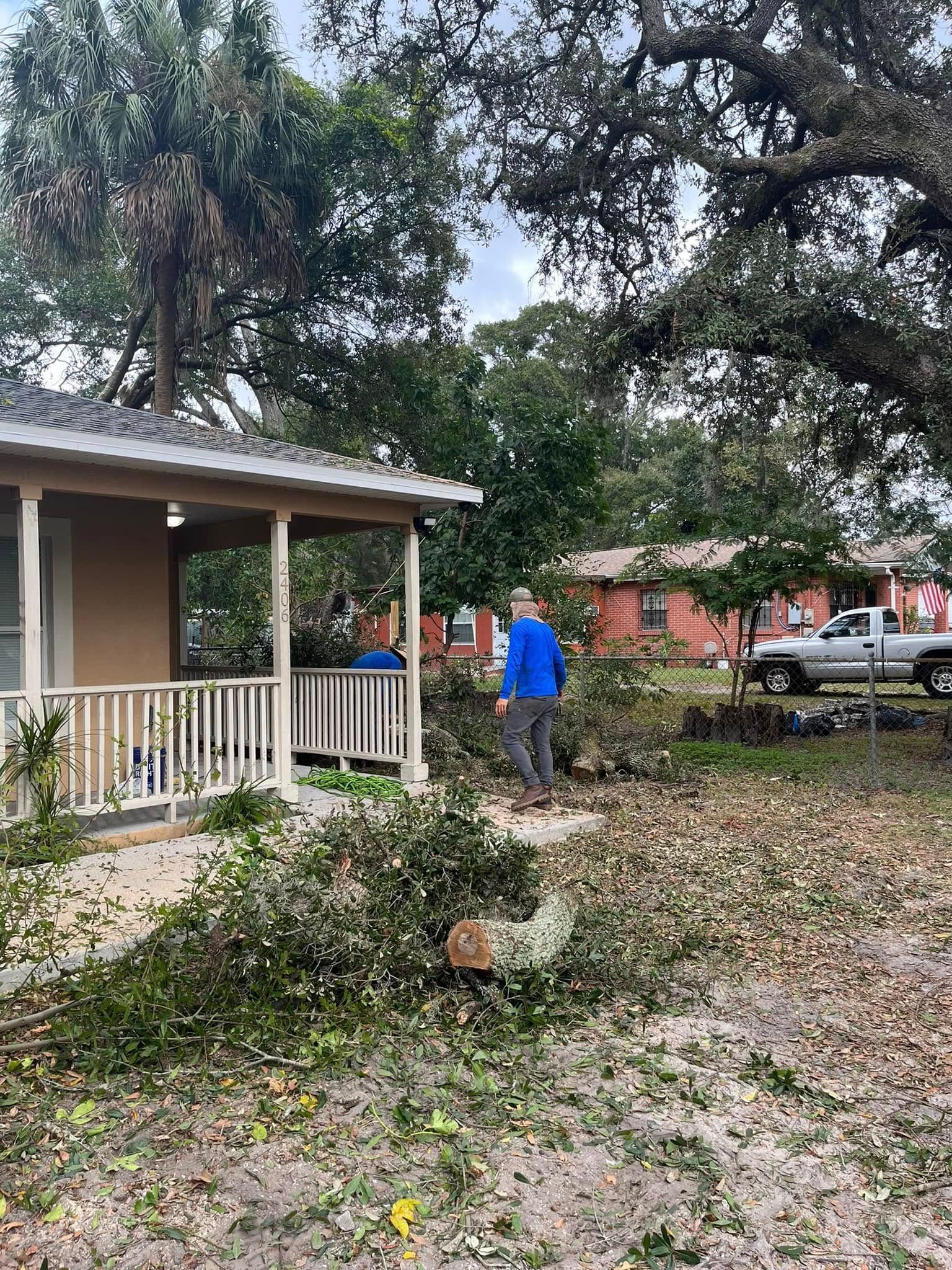 Yard with fallen tree branches, a person in a blue shirt, and a house with a porch after storm damage