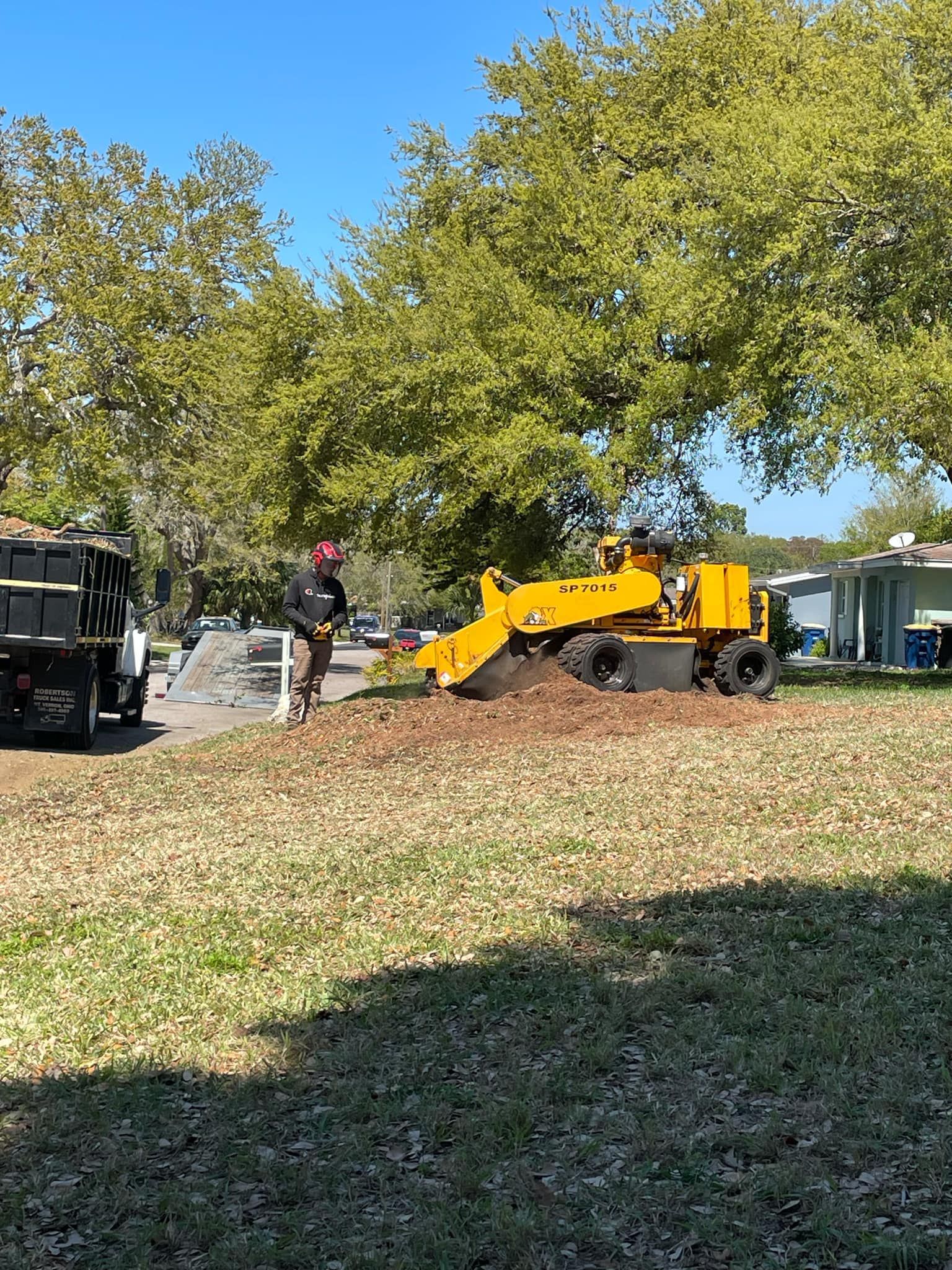 Yellow construction loader moving dirt in a yard with trees and a worker nearby