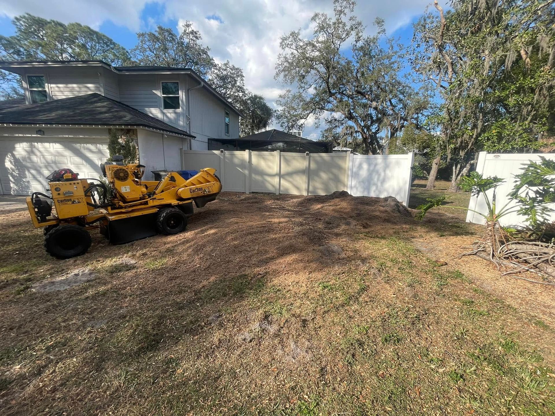 Yellow excavator in a dirt backyard beside a white fence and house under a blue sky Yellow excavator in a dirt backyard beside a white fence and house under a blue sky