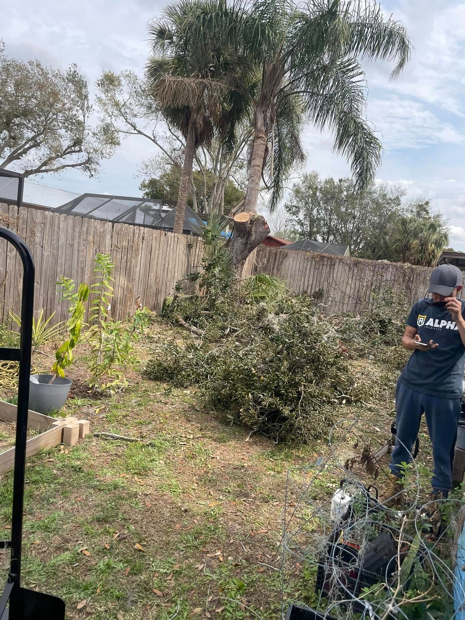 Person in navy work clothes trimming a shrub in a backyard beside a stone wall and trees.