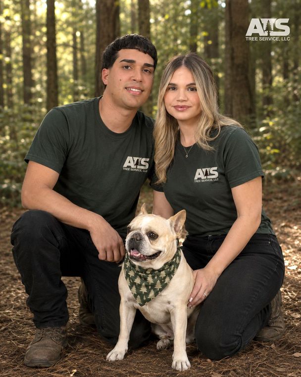 Two people in matching black shirts pose with a small dog in a forest setting.