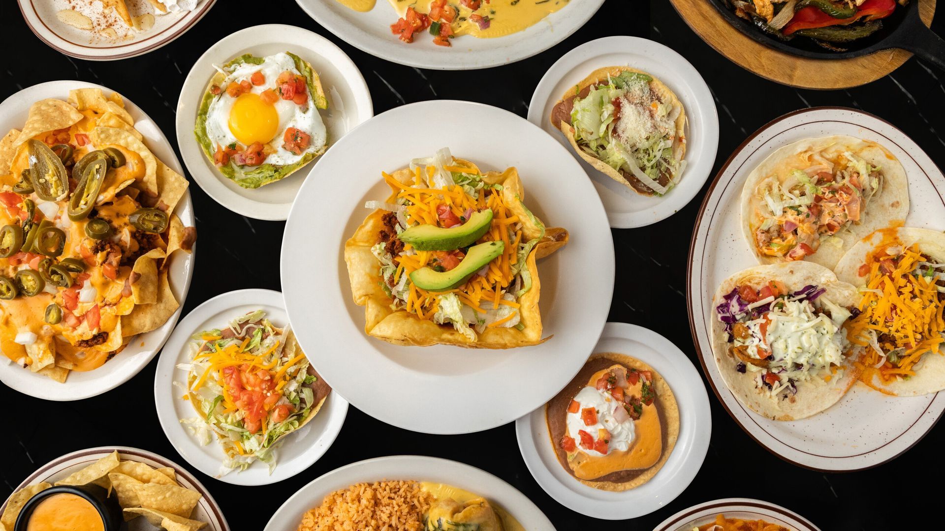Overhead view of various Mexican dishes including toastadas, nachos, taco salad, and soft tacos on a black surface.