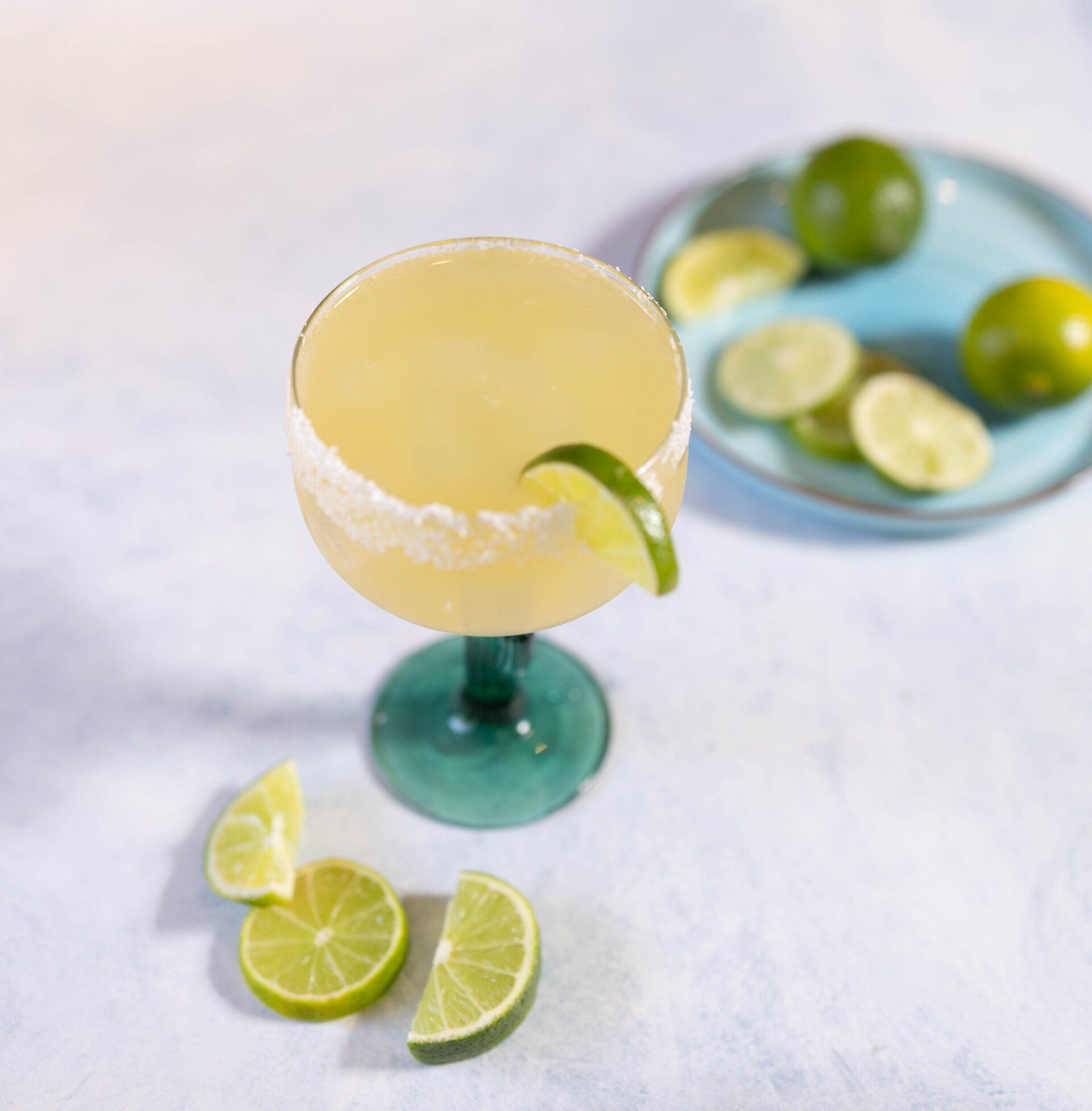 House Margarita in a salt-rimmed glass, lime slice garnish, surrounded by lime wedges and whole limes on a blue plate.