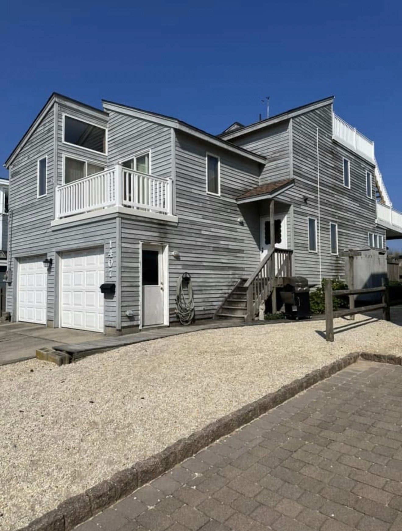 Two-story, weathered gray house with white garage doors and a balcony on a gravel lot.