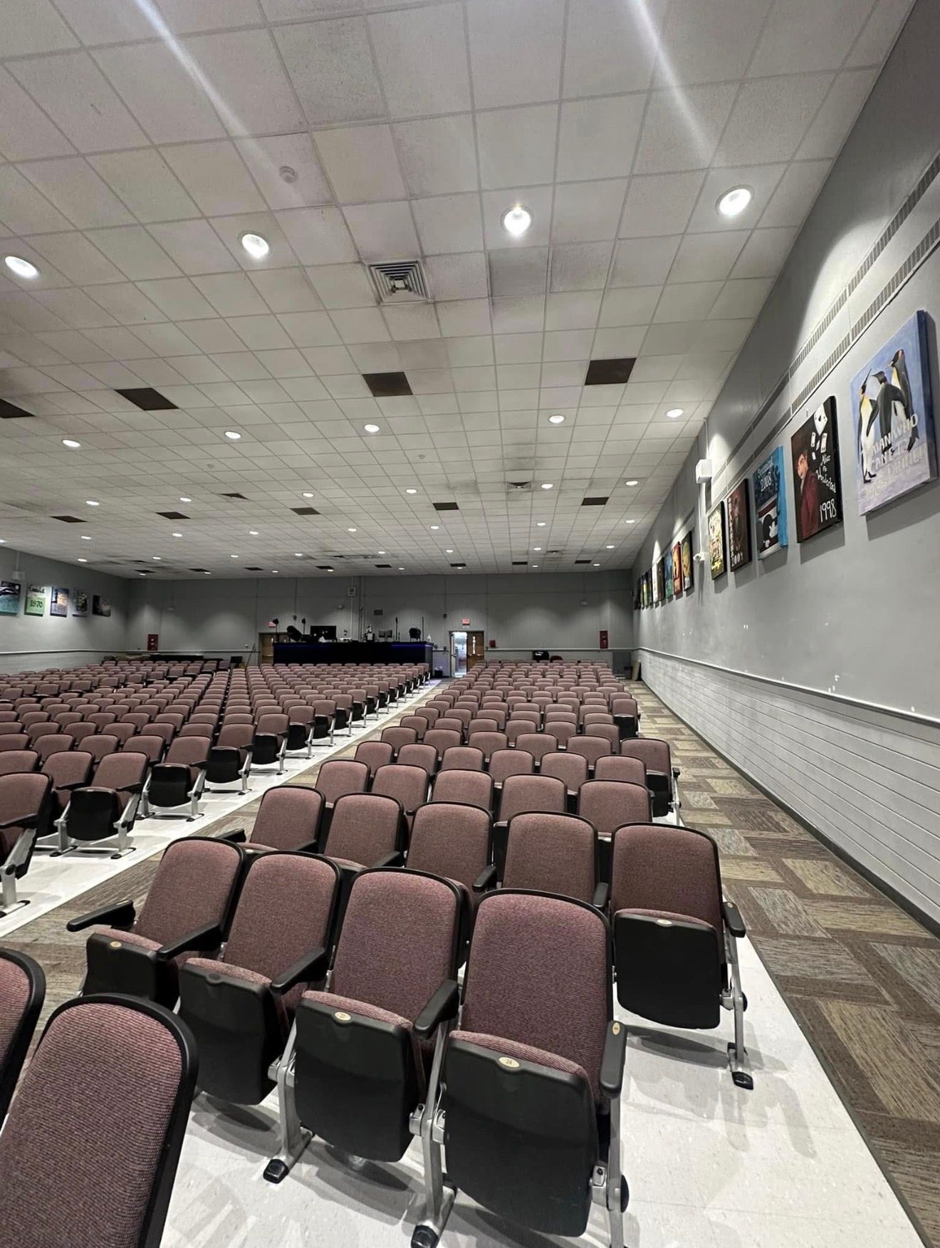 Rows of empty maroon auditorium seats facing a stage. Portraits line the right wall.