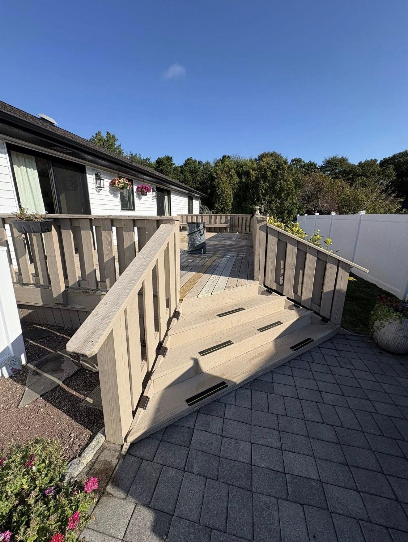 Deck with ramp, gray pavers, beige railing, and white building in a sunny setting.