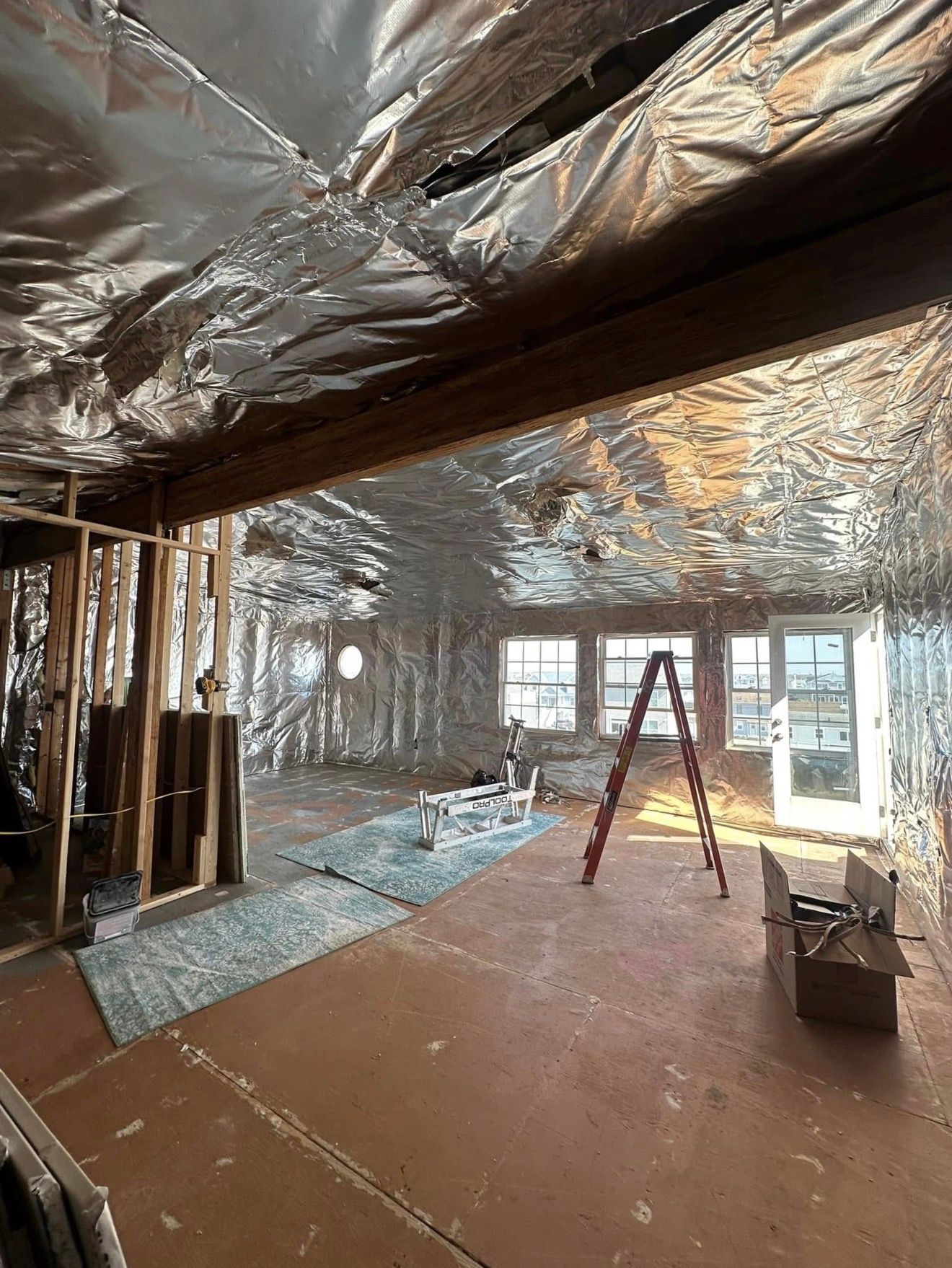 Interior of a room under construction with reflective insulation on walls and ceiling, ladder, and windows.