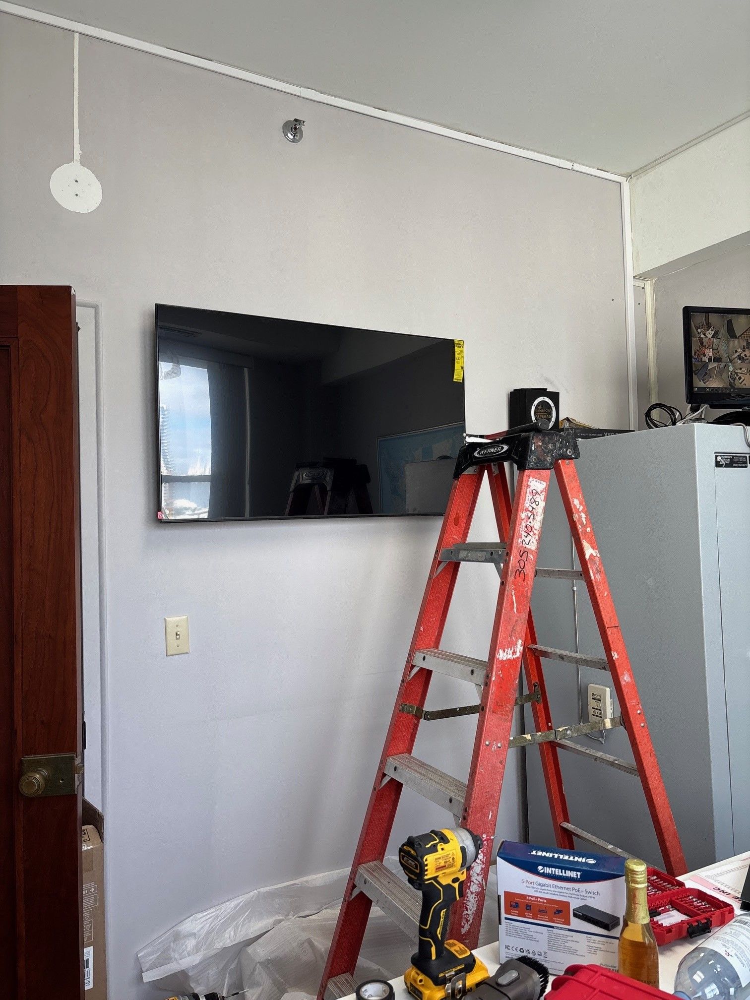 A red ladder stands by a wall-mounted TV. A drill, paint supplies, and electrical wire are visible.