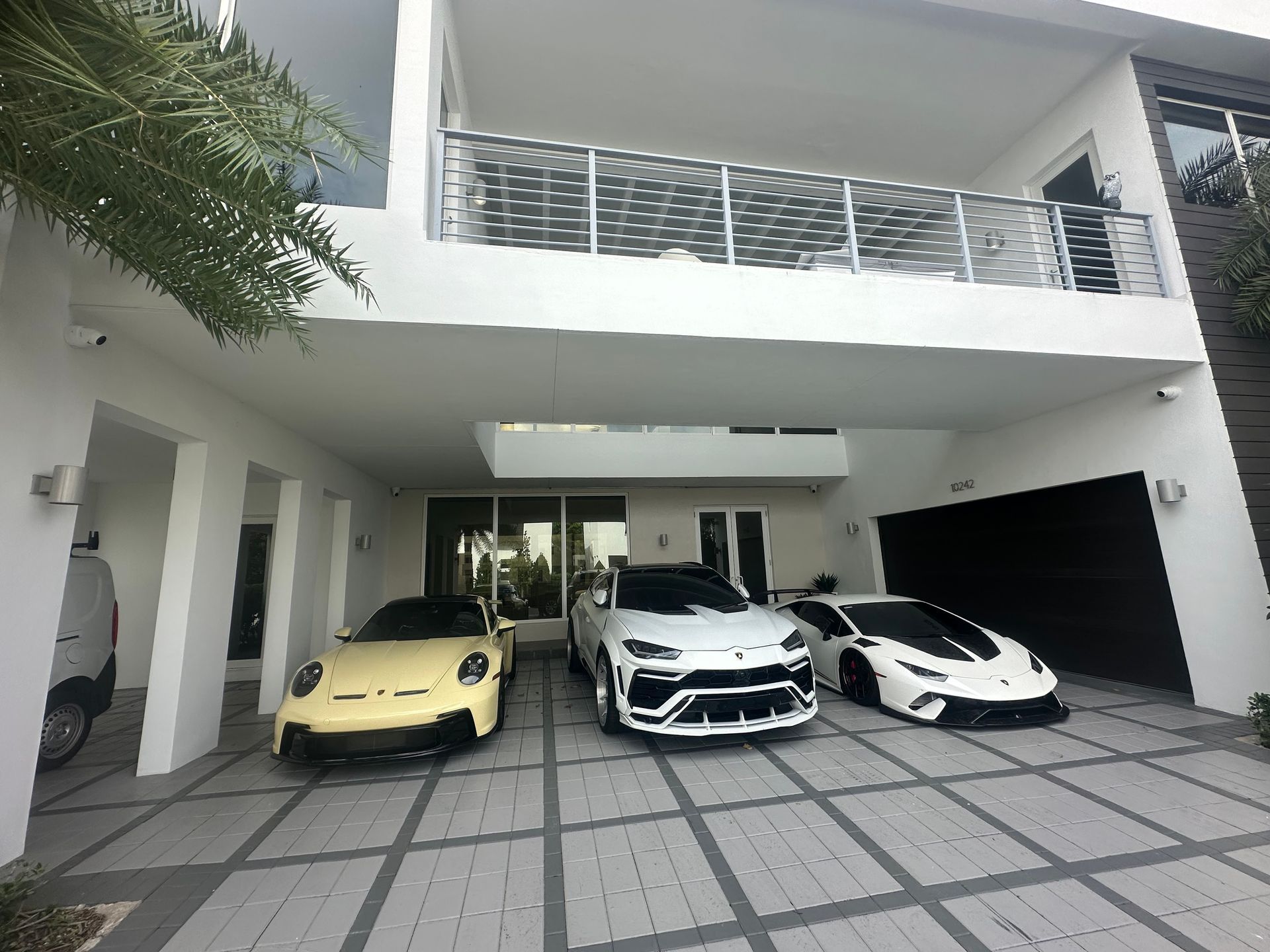 A luxury home with three sports cars parked under a covered area, including a yellow Porsche.
