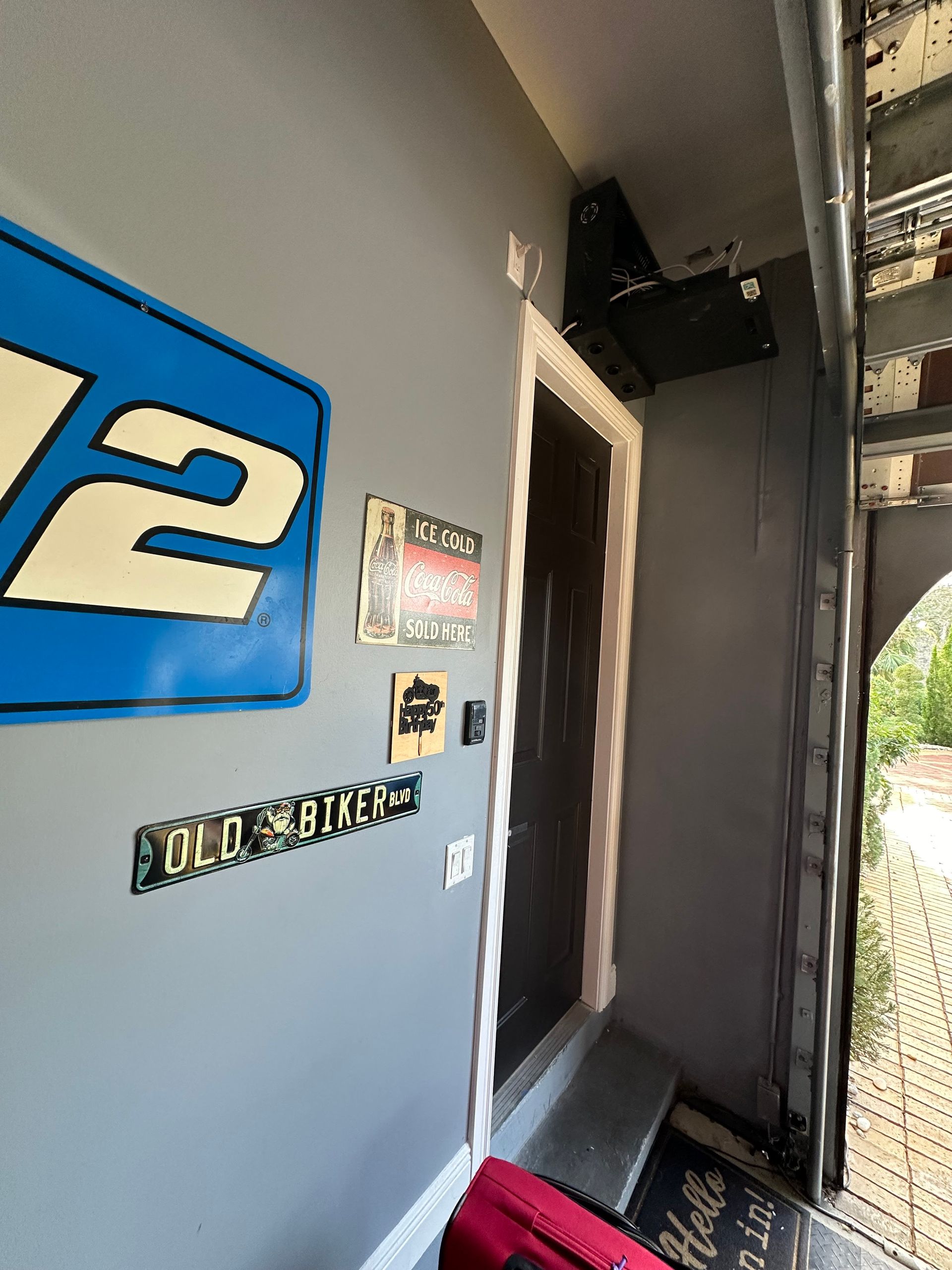 Entryway with a blue sign, black door, and a security camera. Gray wall and a welcome mat.