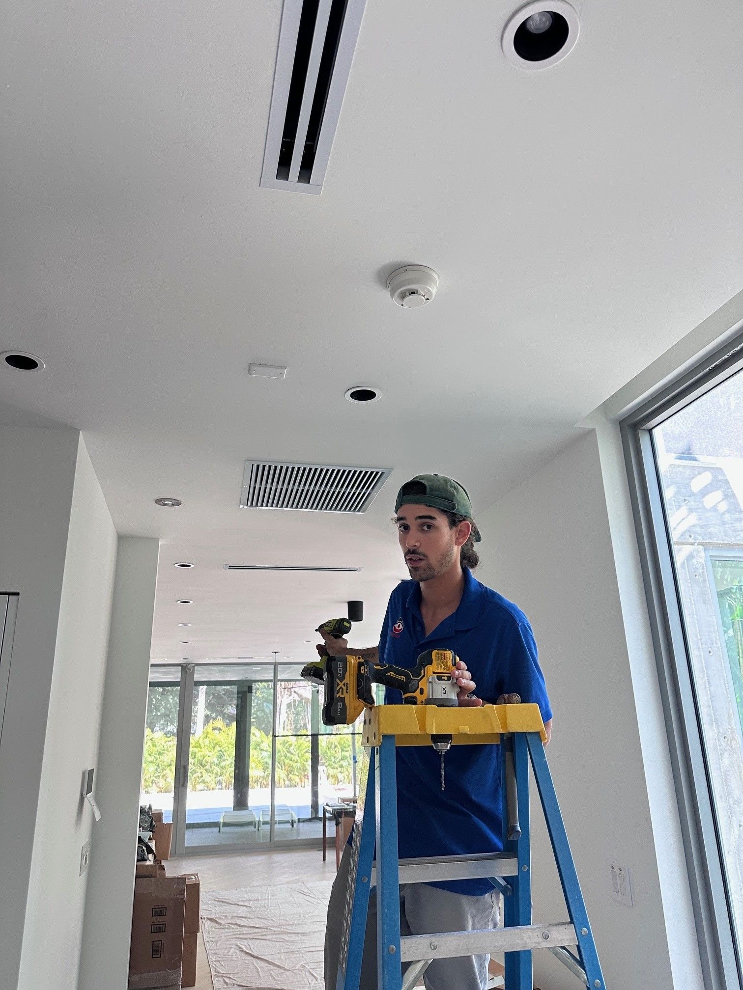 Man on ladder installing light fixture in white-walled room; ceiling vents, windows.