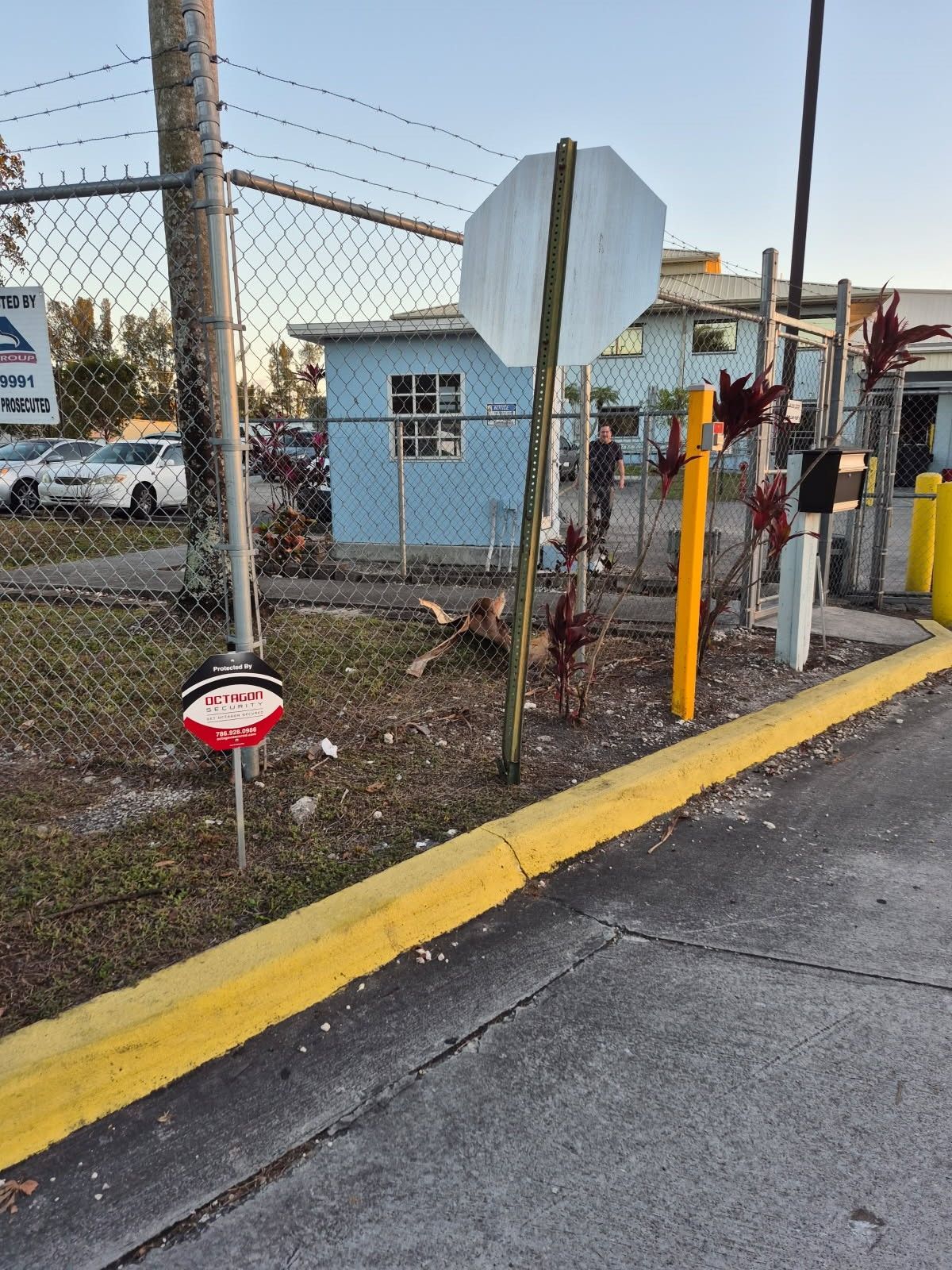 Chain-link fence with a stop sign, security sign, and yellow curb alongside a driveway; a blue building is in the background.