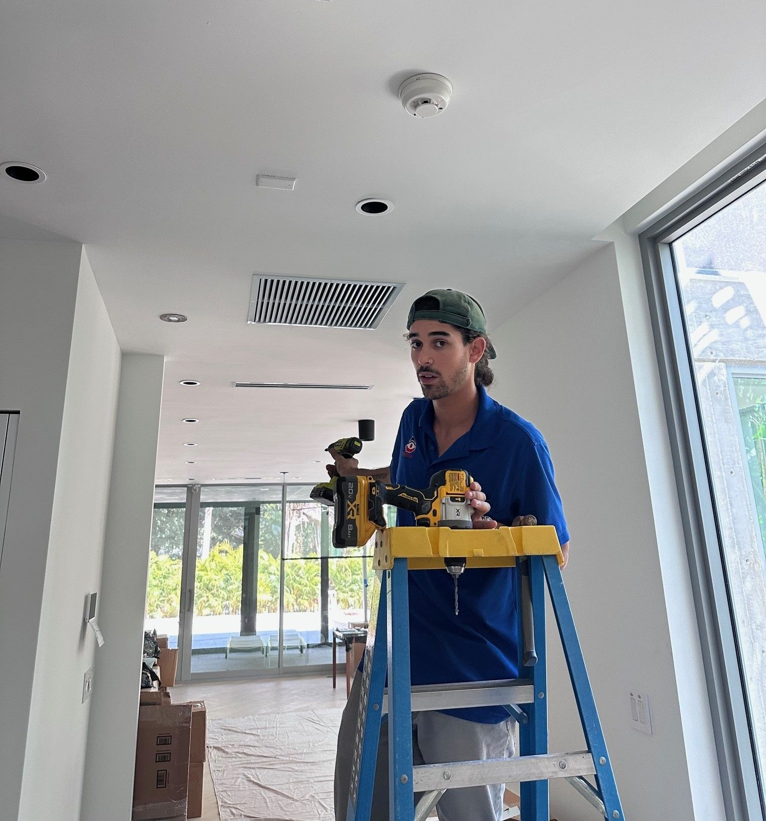 Man on ladder installing equipment on a ceiling indoors. He wears blue shirt, green hat, and looks toward the camera.