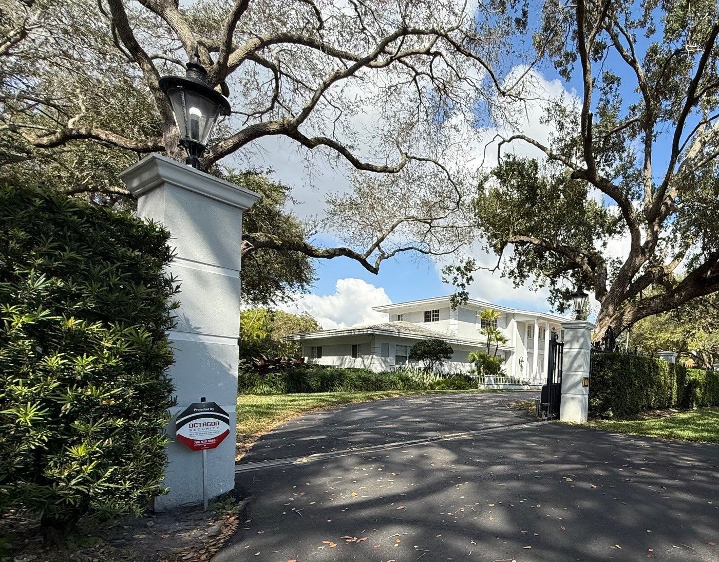 Gated white mansion entrance with a driveway, mature trees, and a security system on a pillar.