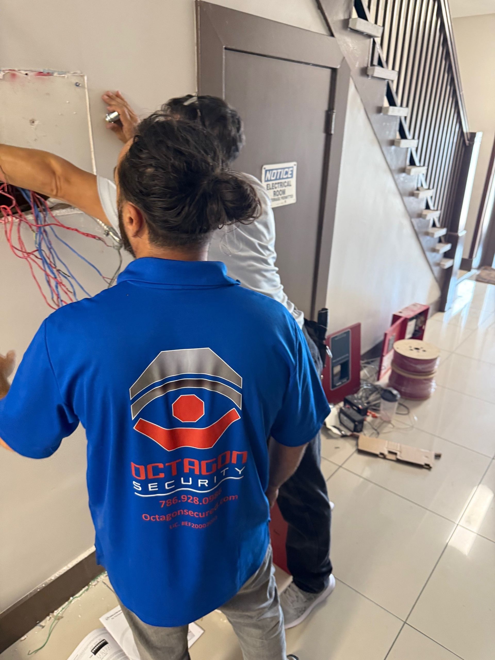 Two people installing electrical wiring on a wall indoors. One wears a blue Octagon Security shirt.