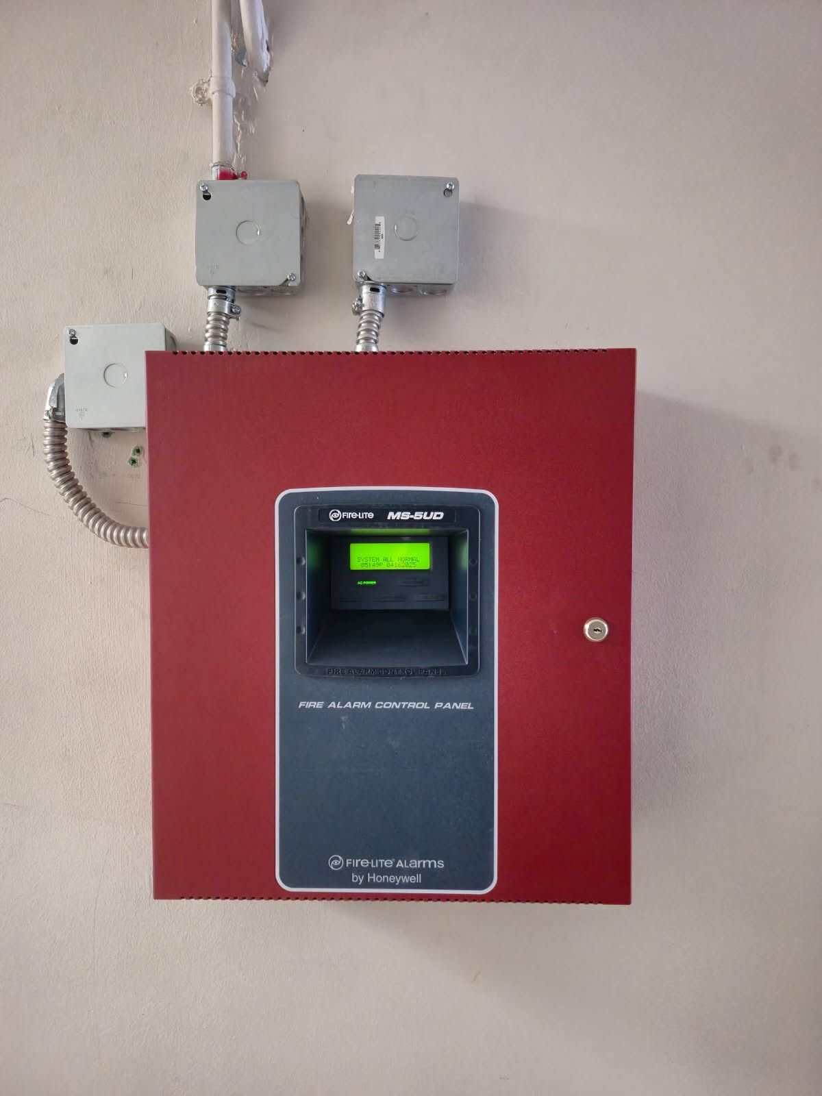 Red fire alarm control panel mounted on a beige wall with electrical boxes above.