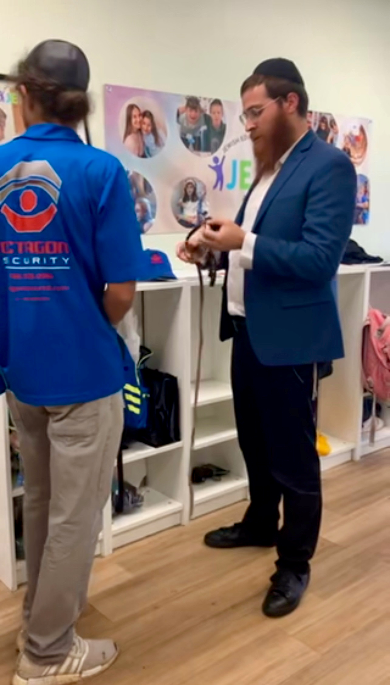 Man in blue blazer examines object; another man in security shirt stands nearby. White shelves in background.
