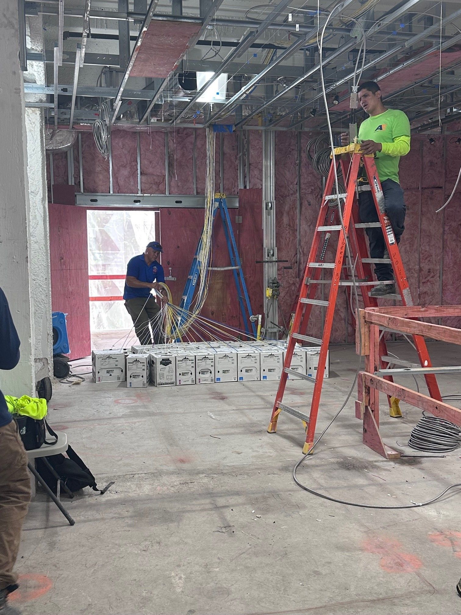 Construction workers wiring electrical in a building under renovation. One on a ladder, another at the electrical panel. 