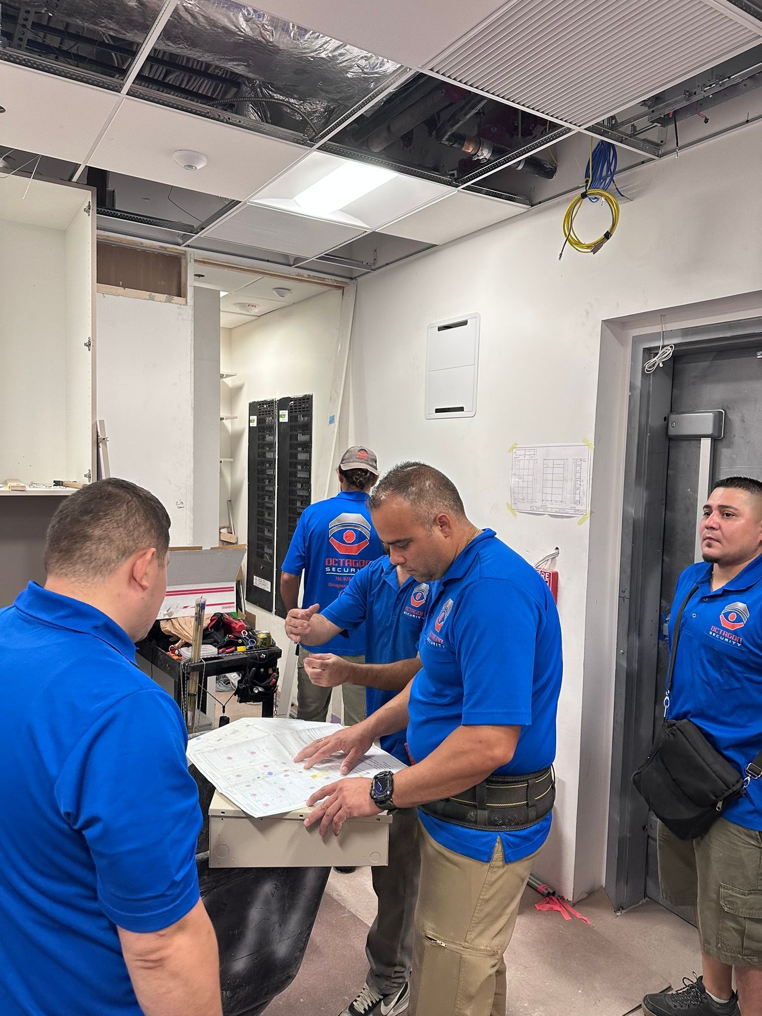 Men in blue shirts reviewing blueprints in a room under construction with exposed ceiling.