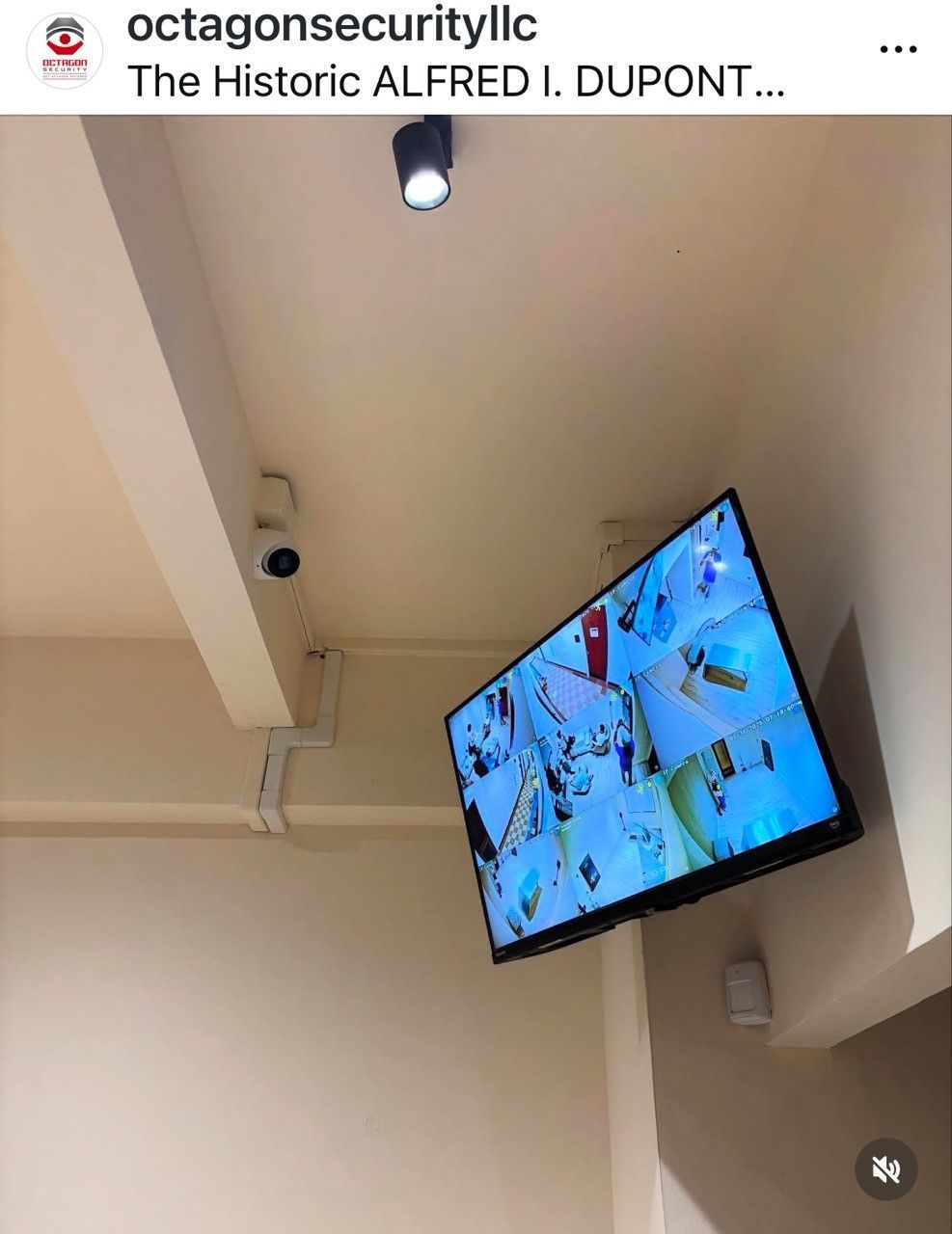 Ceiling-mounted security cameras and monitor, showing security footage, in a building with white walls and a light fixture.