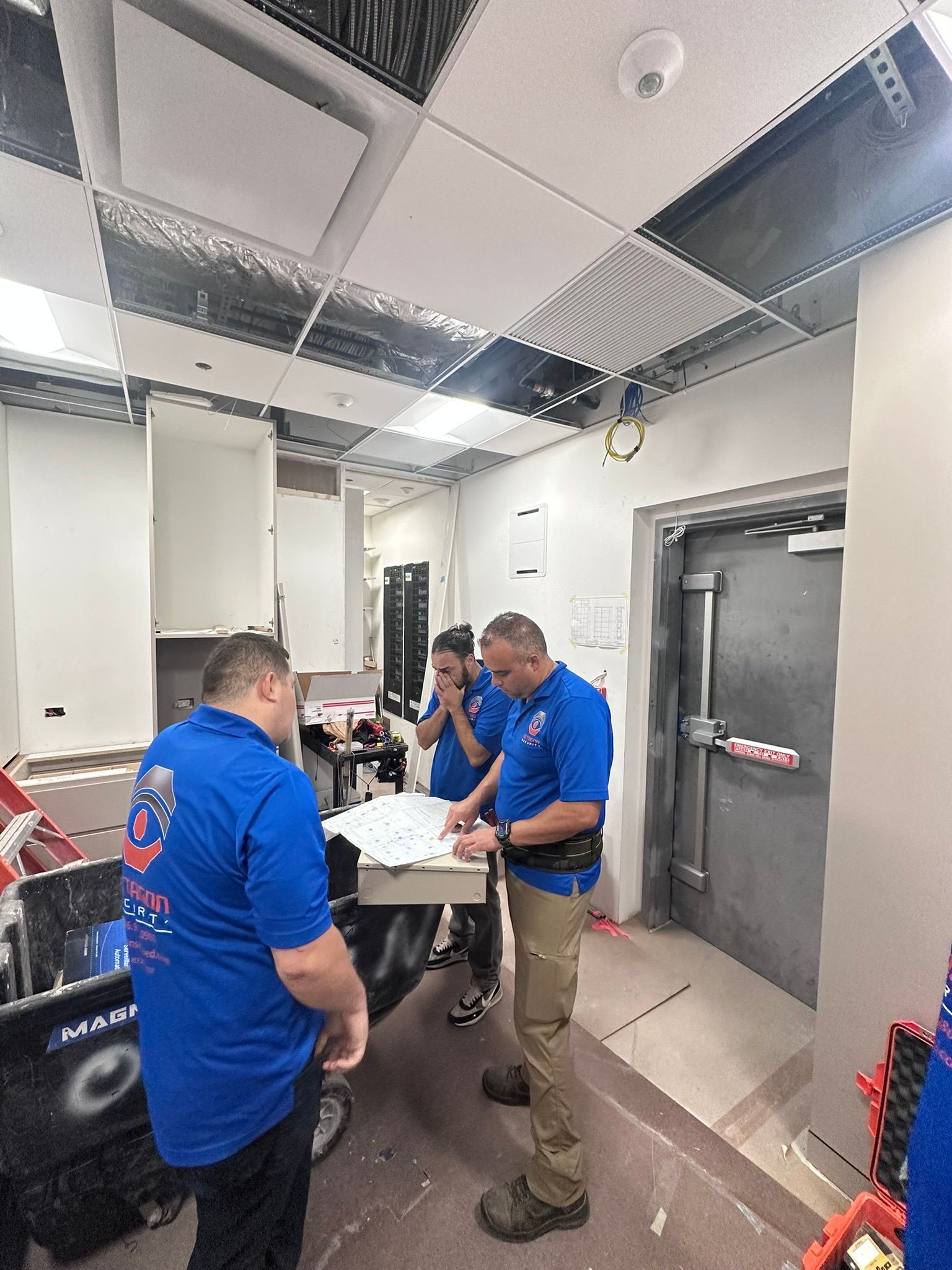 Three people in blue shirts looking at papers in a room with exposed ceiling infrastructure.