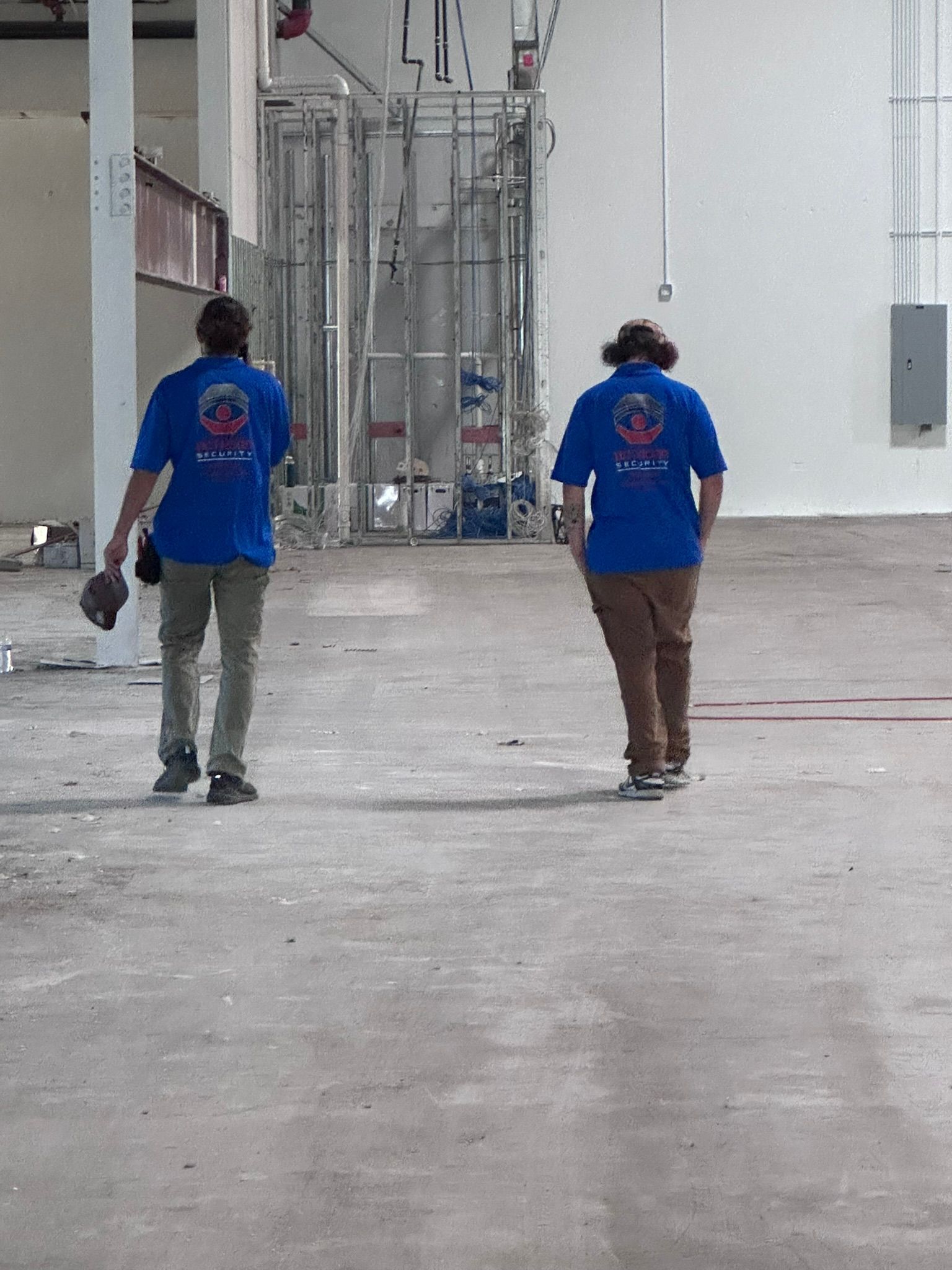 Two people in blue shirts walk away, inside a building. Grey floor, industrial background.