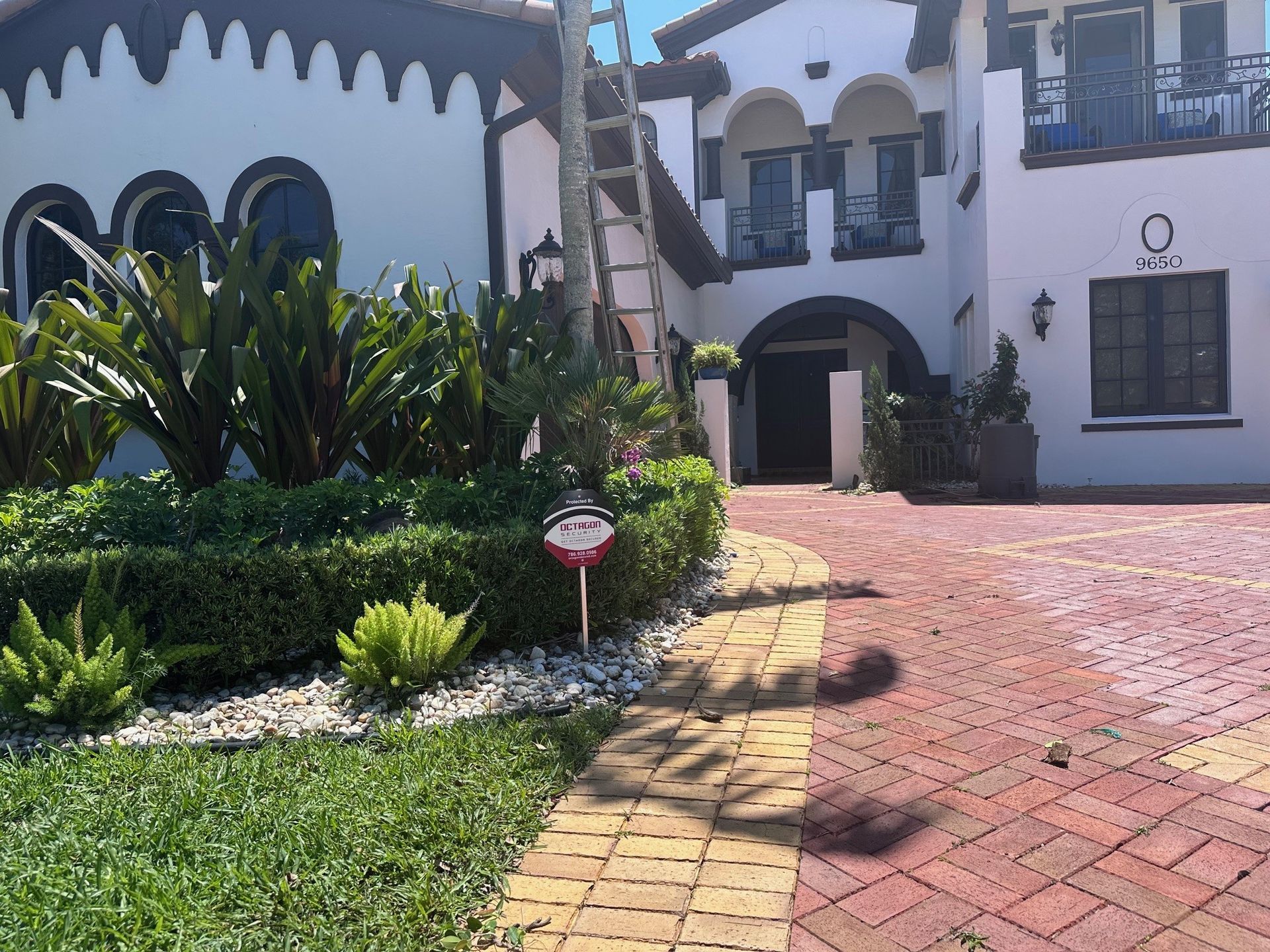 Two-story stucco house with arched doorways, red brick driveway, ladder, and manicured landscaping.