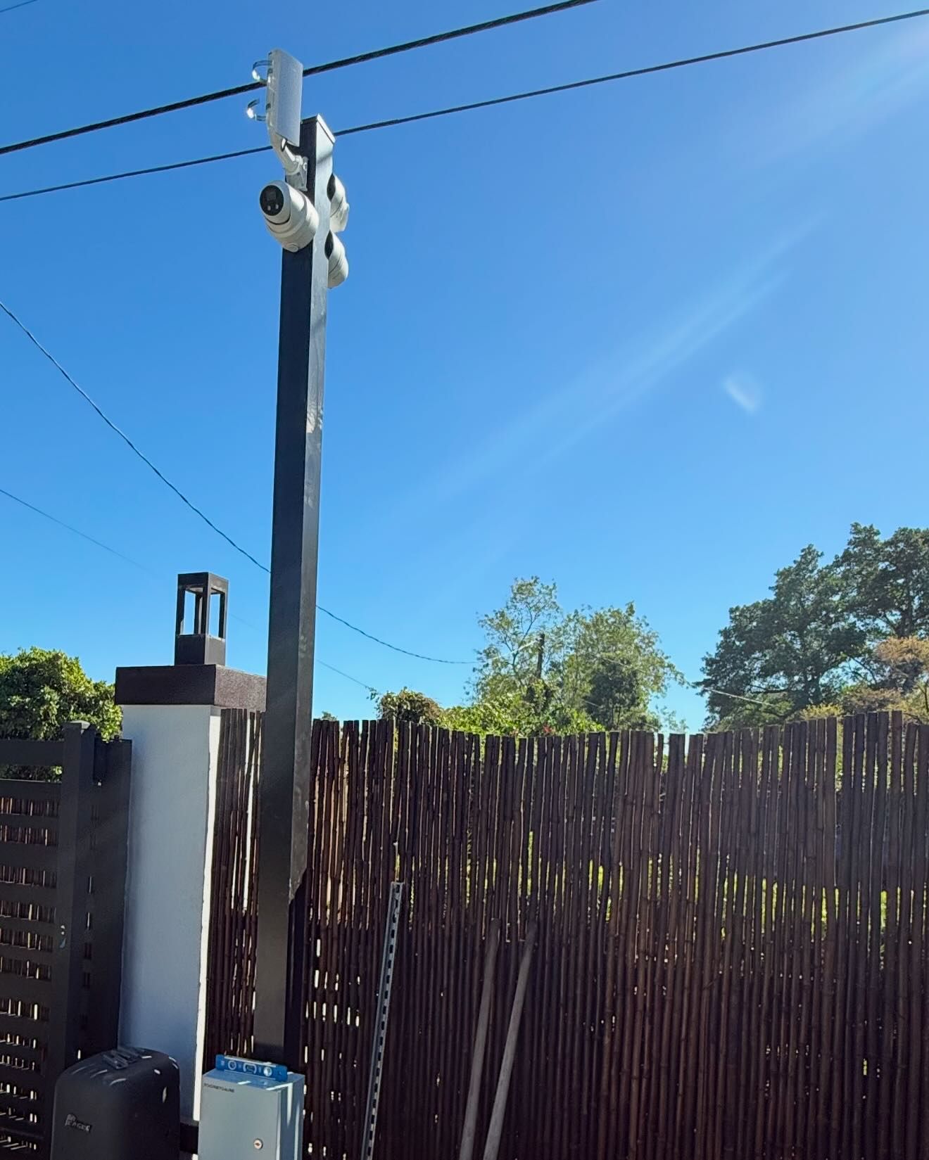 Security cameras on a tall pole, mounted beside a fence, against a blue sky.