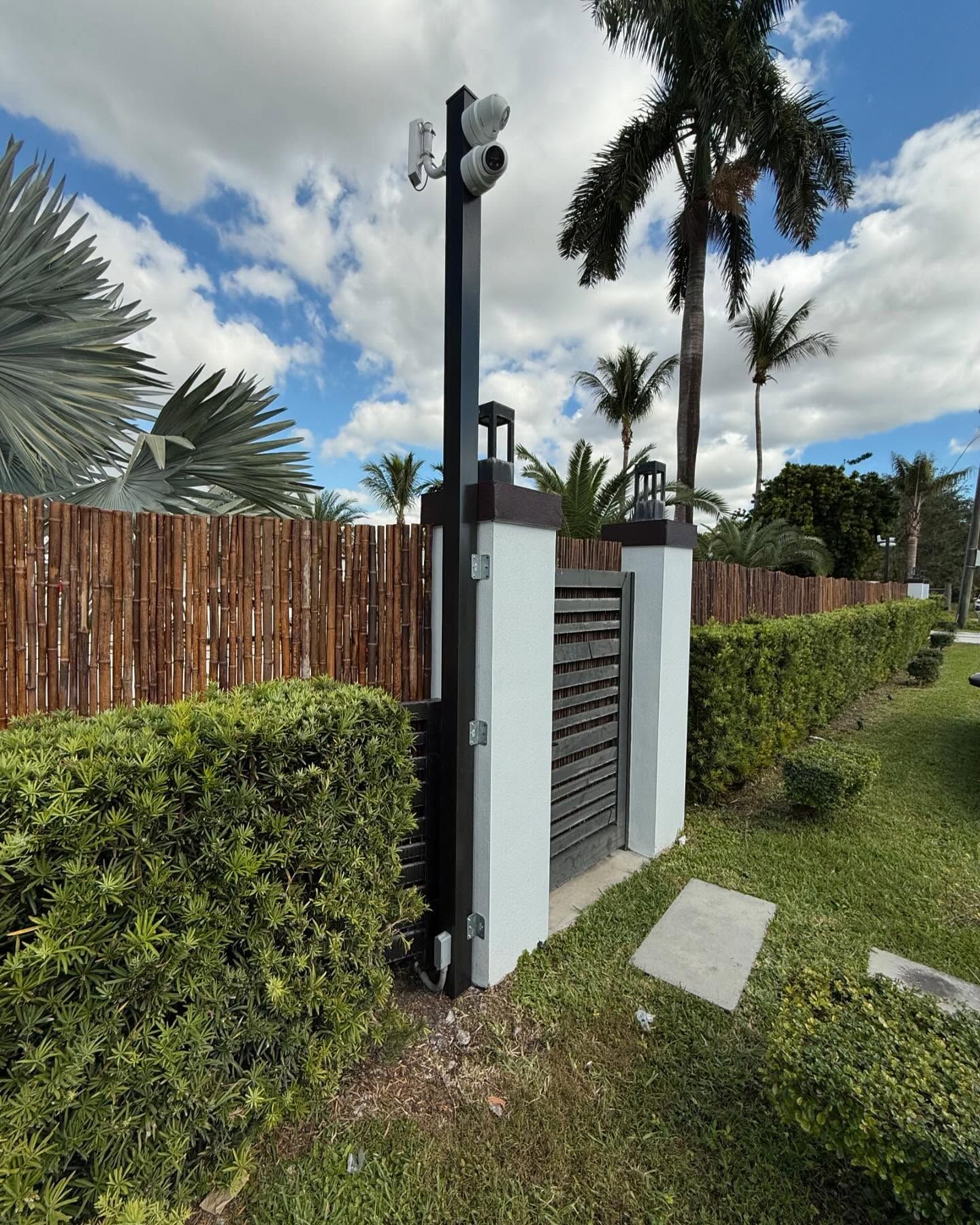 Black gate with security cameras, next to a brown wooden fence, green bushes, and palm trees under a cloudy blue sky.