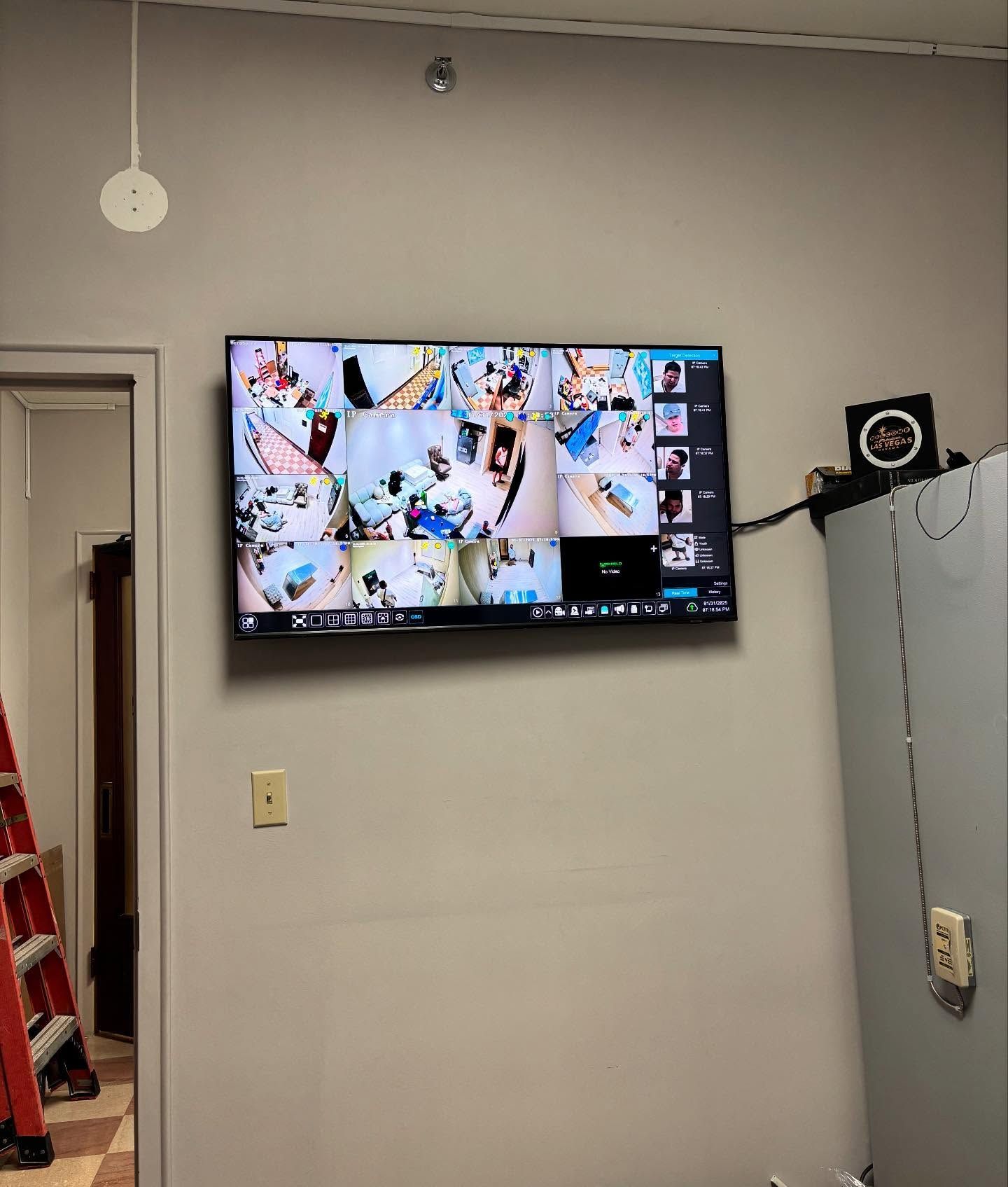 Security camera monitor on a gray wall, showing multiple camera views. A clock is nearby, plus door and ladder.