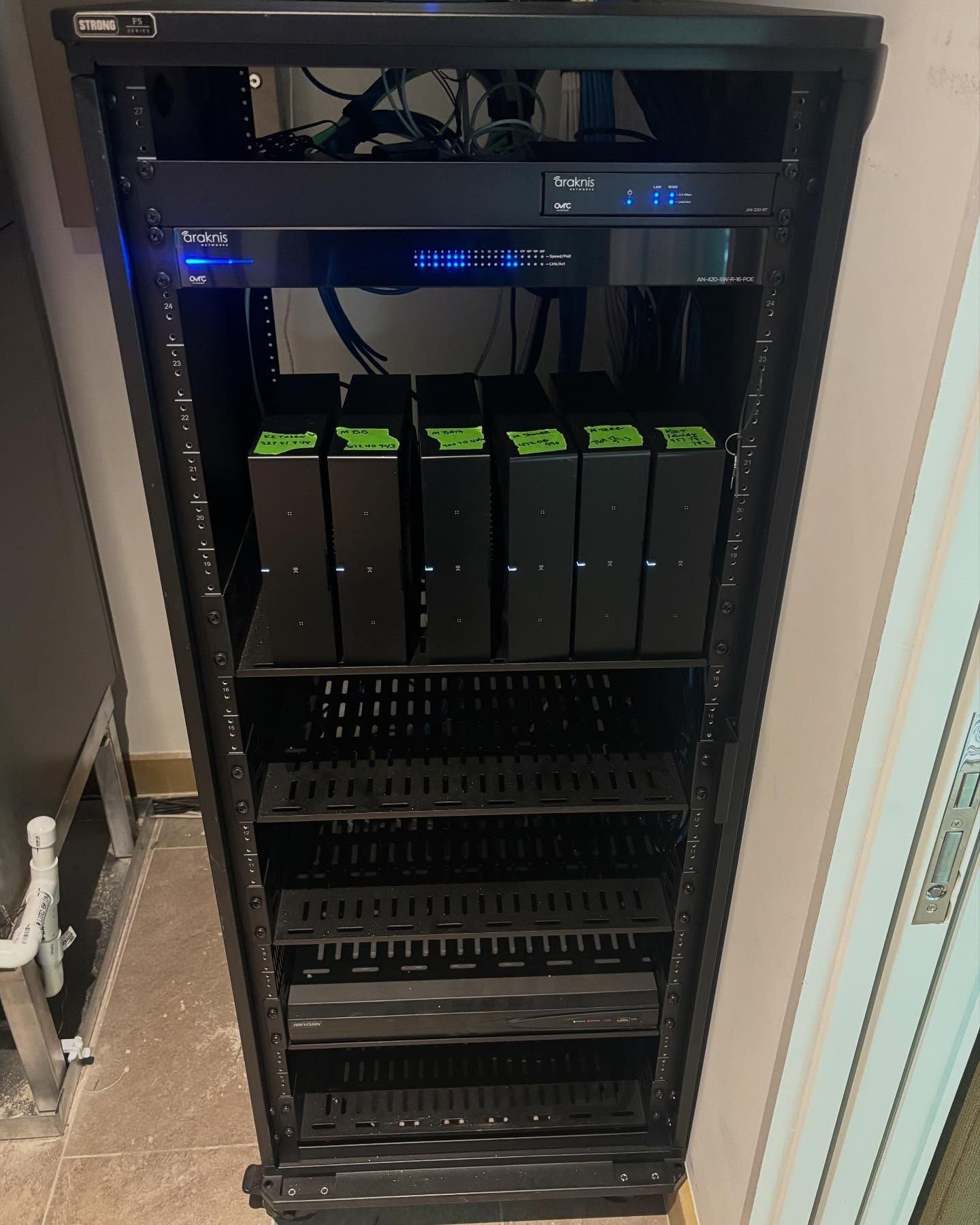 Black server rack with multiple black devices and green accents. Located in a room.