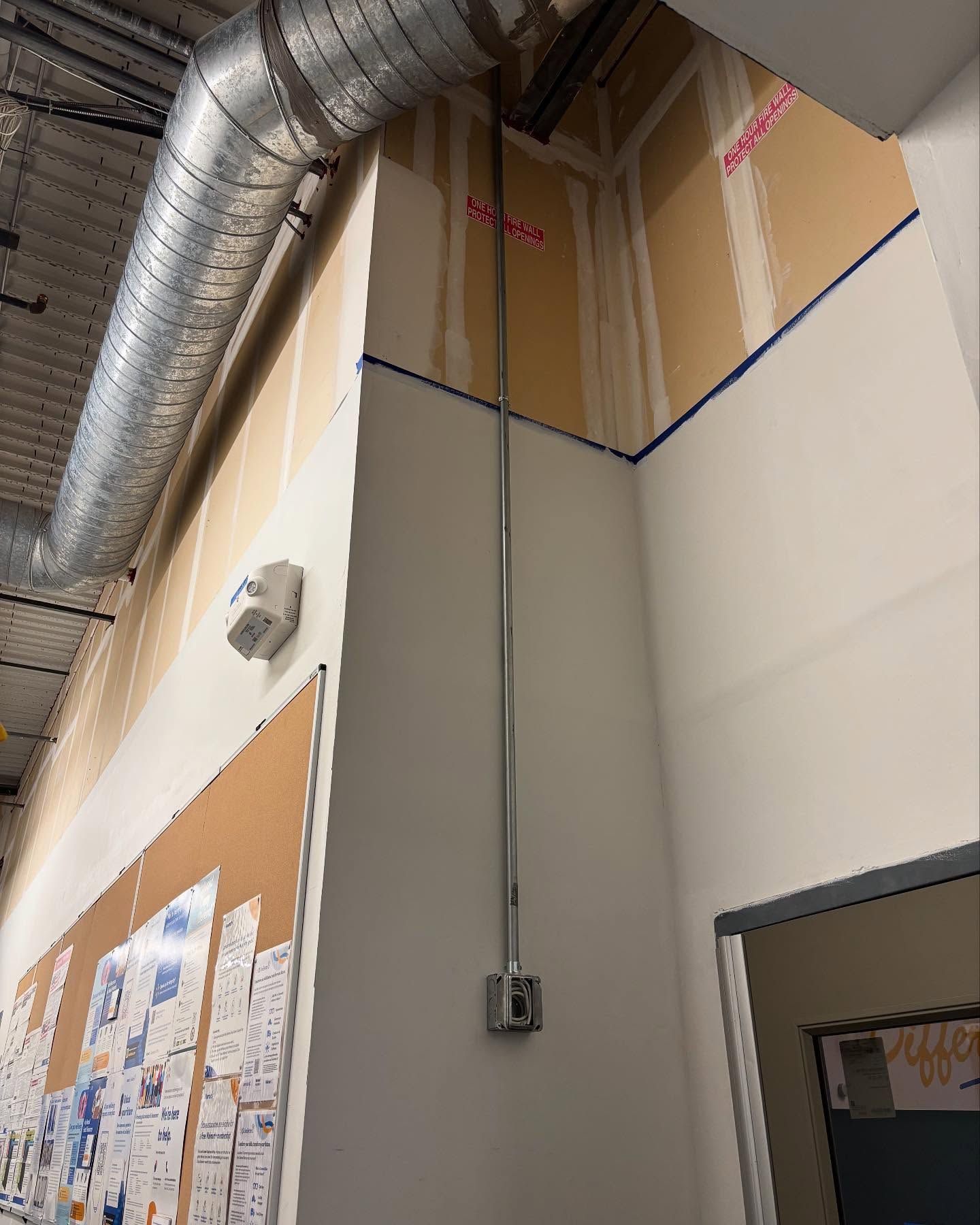 Silver conduit runs down a white wall in an industrial setting, with a vent and bulletin board visible.